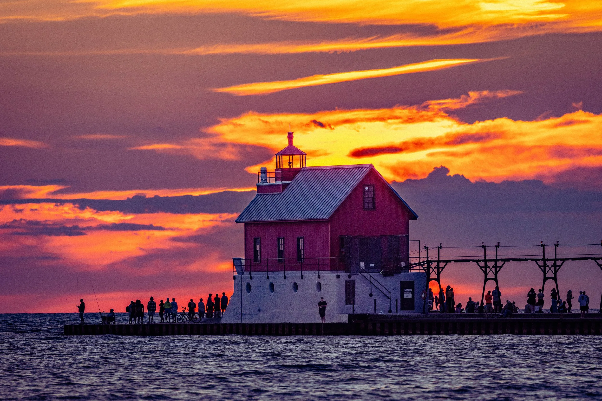 A lighthouse on a pier during sunset with a colorful sky, and people gathered along the edge of the pier.