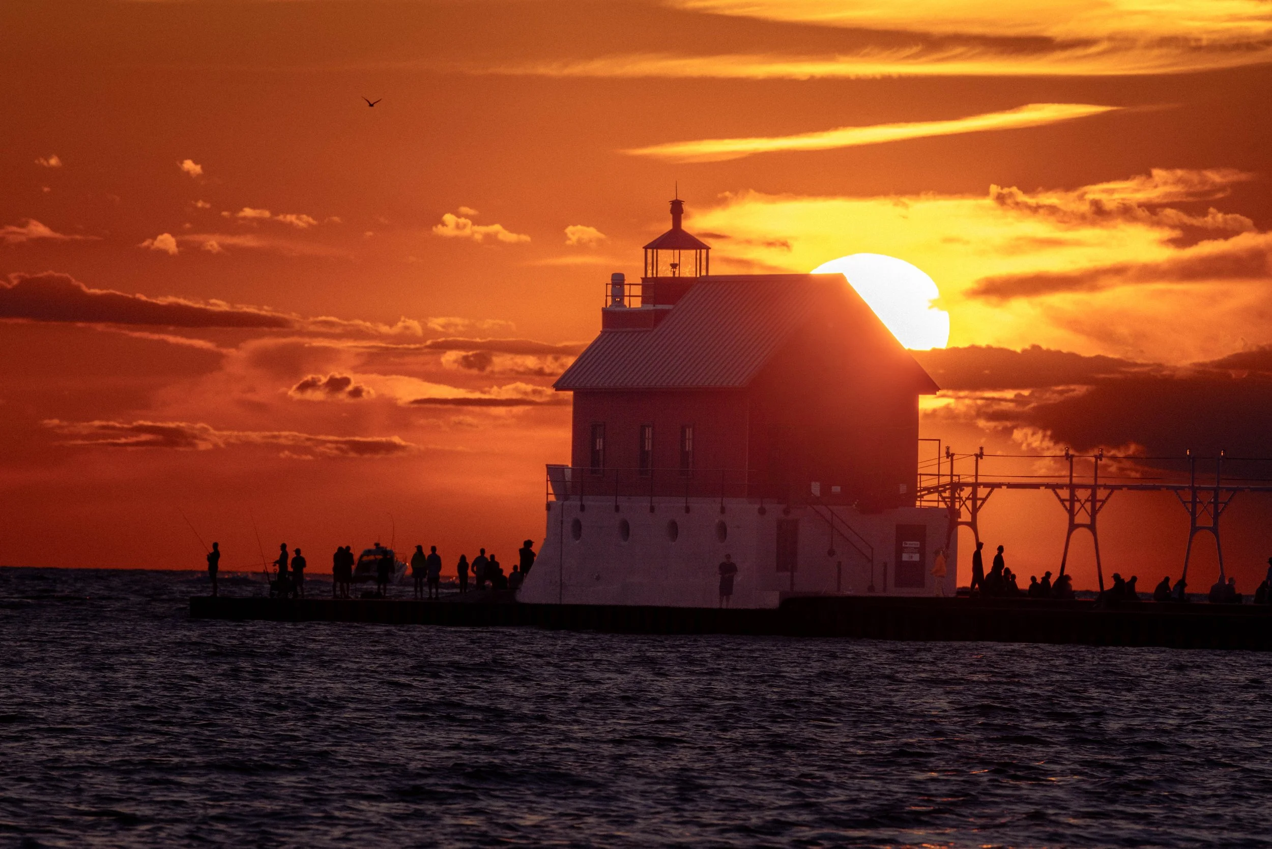 Sunset over the ocean with a lighthouse in the foreground, silhouetted people on the pier, and a flying bird in the sky.