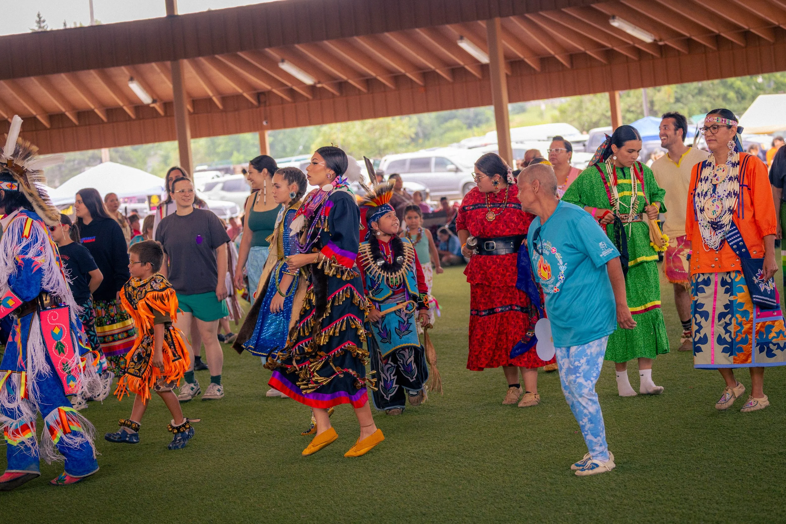 People dressed in traditional Native American clothing at a cultural gathering or festival, walking and interacting under a pavilion with parked vehicles in the background.