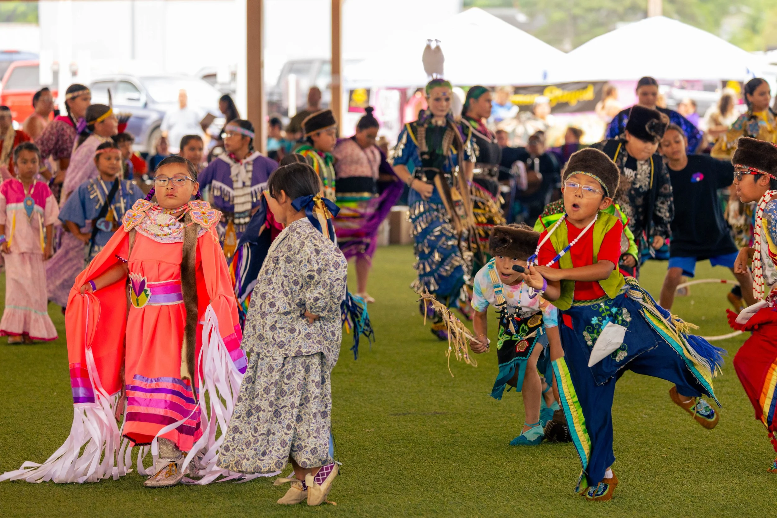 Children and adults in colorful traditional Native American clothing participating in a cultural dance at an outdoor festival under a large white tent.