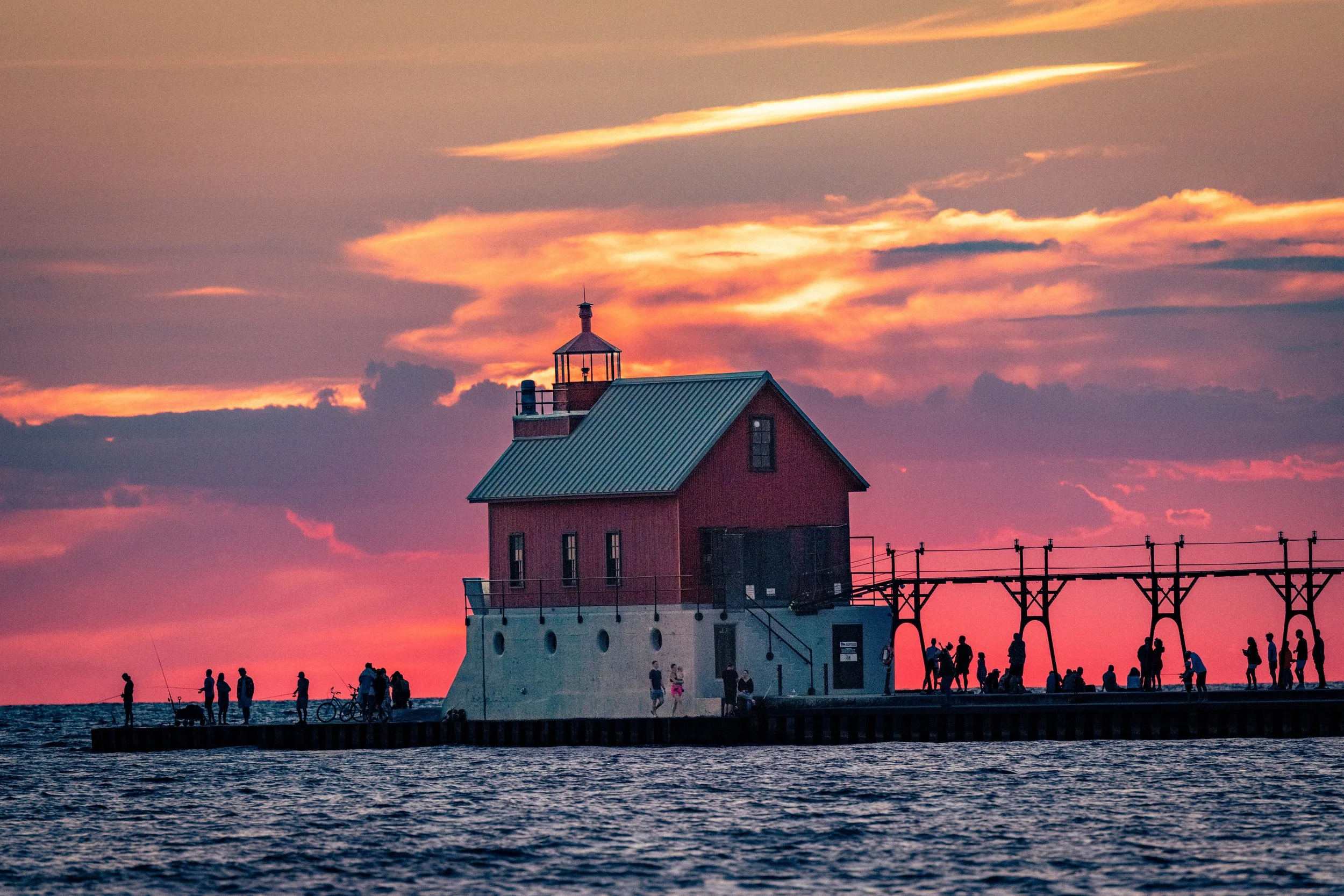 A lighthouse on a pier during sunset with people walking and sitting nearby, the sky filled with pink, orange, and purple clouds.