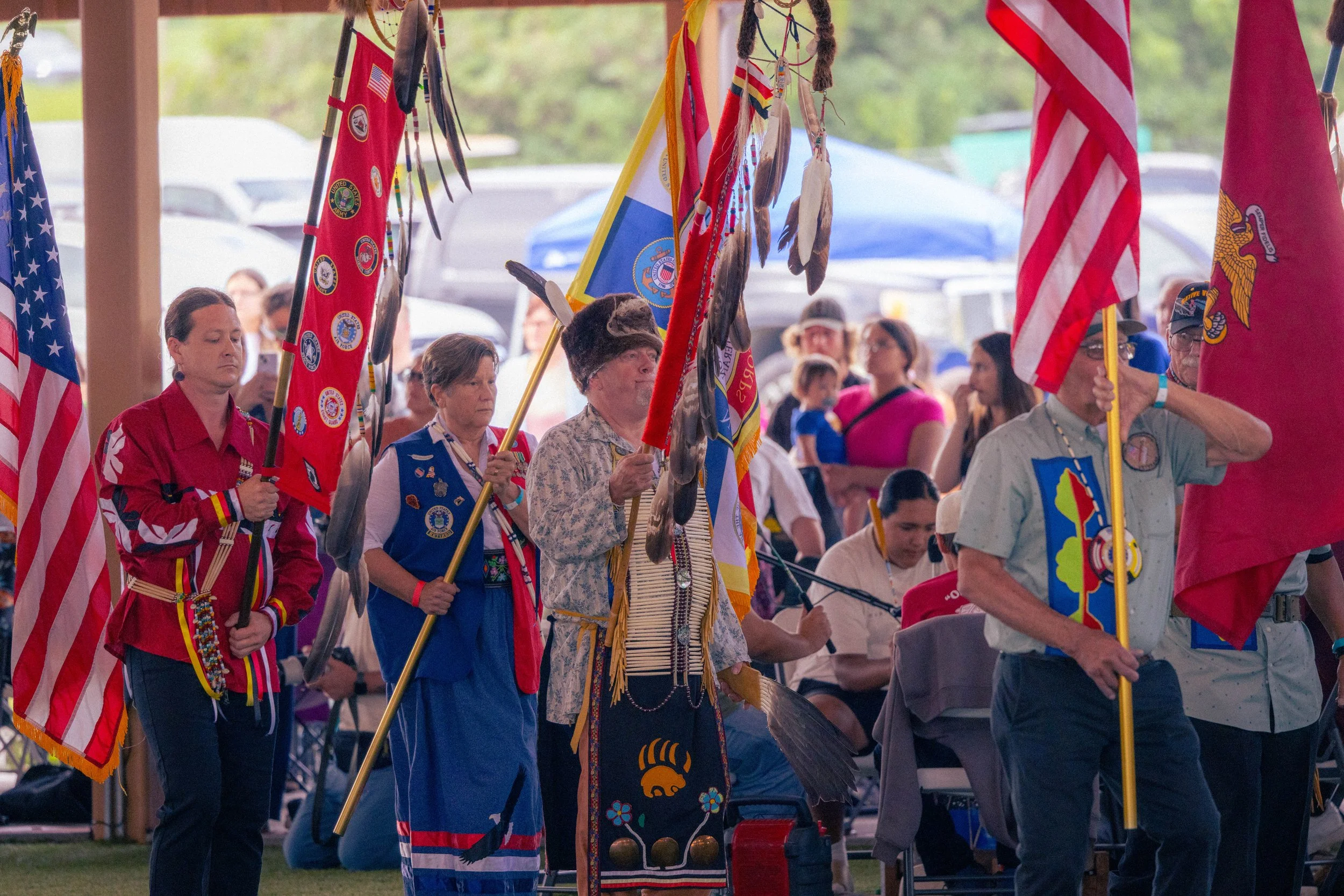 People participating in a Native American cultural ceremony, holding flags and traditional regalia, with other attendees in the background.