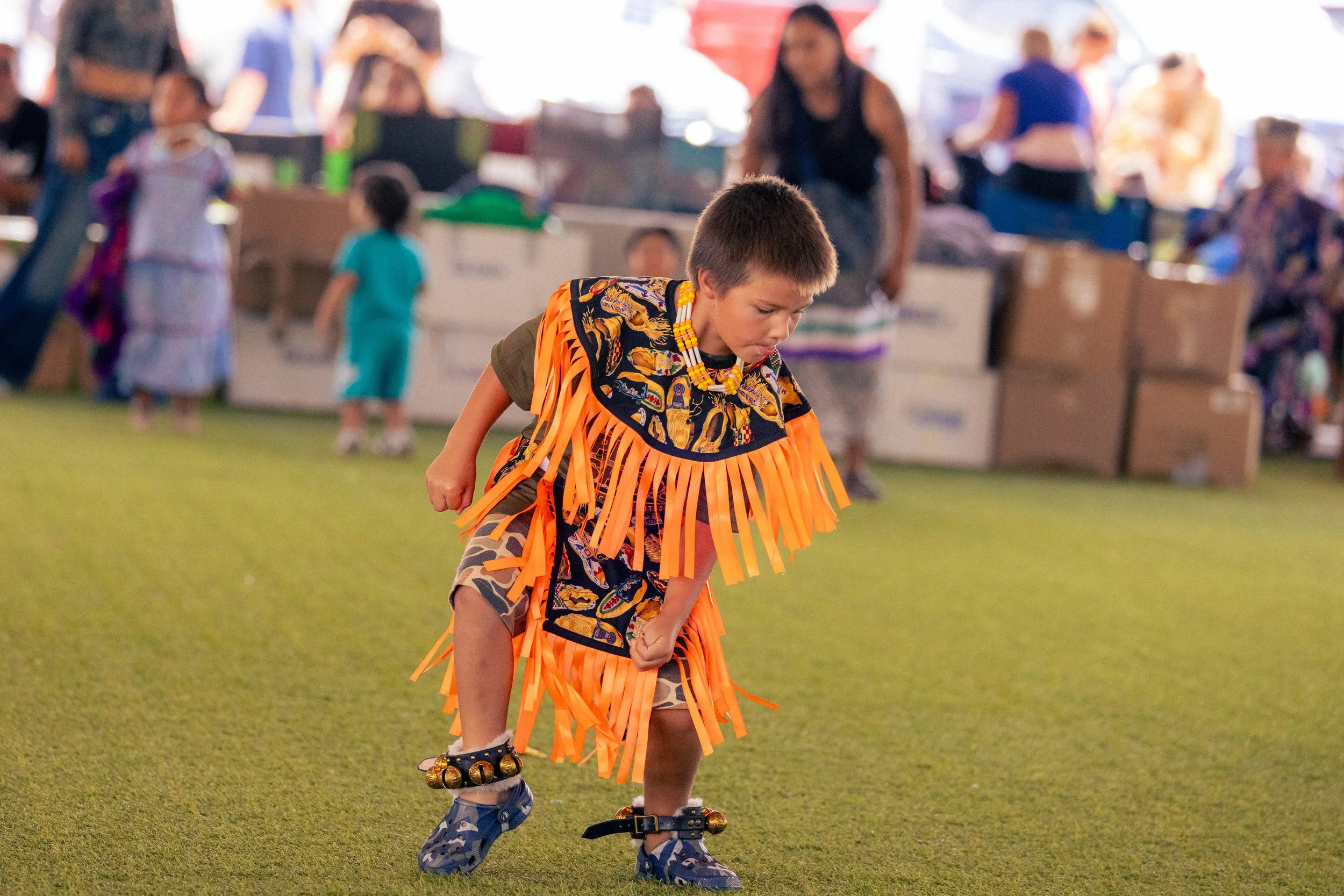 Young boy dressed in traditional indigenous costume with orange fringe, patterned shirt, and accessories, performing a dance at an outdoor event with other participants and spectators in the background.