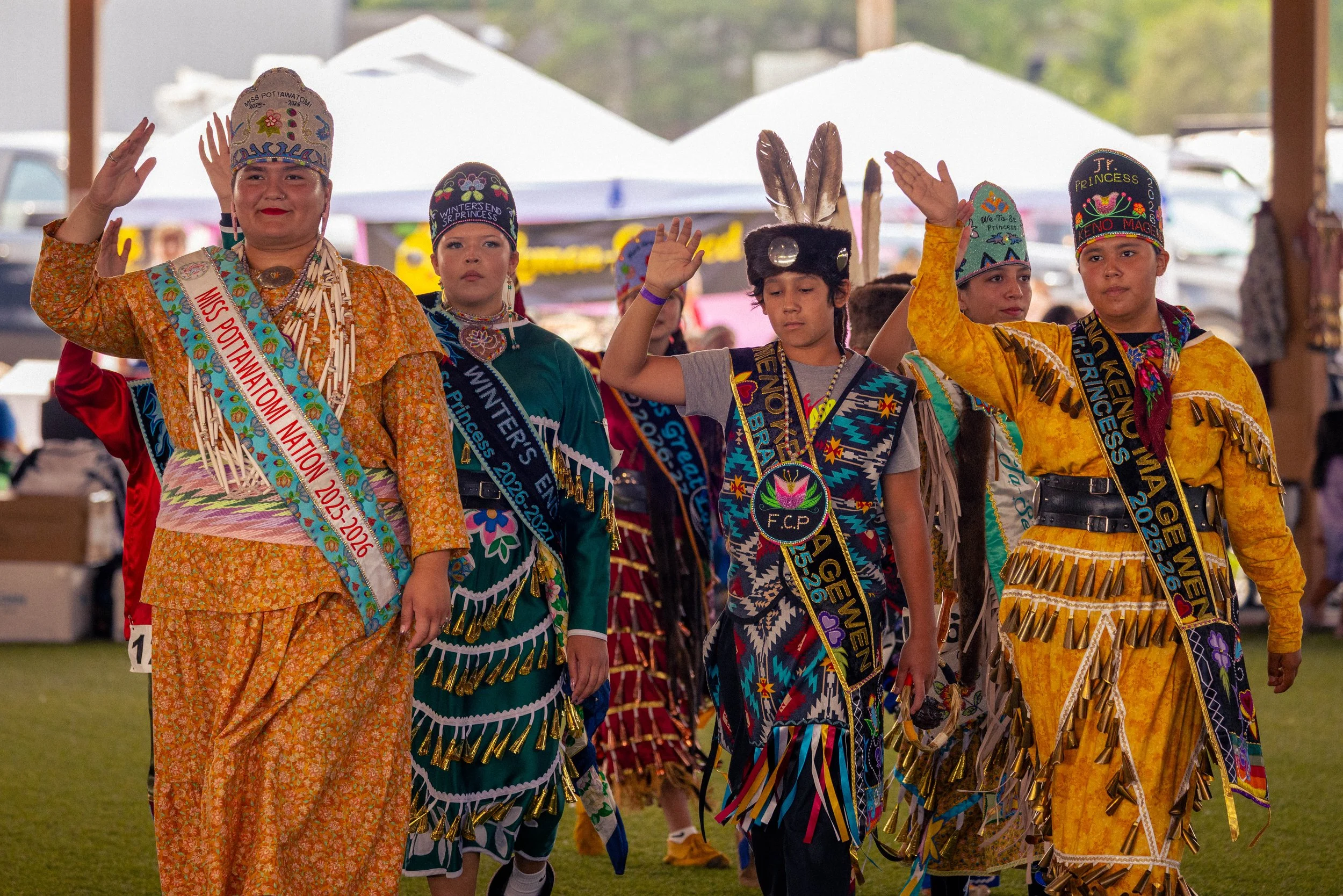 Group of Indigenous women and children dressed in traditional regalia participating in a cultural event or ceremony, with some wearing sashes and hats.