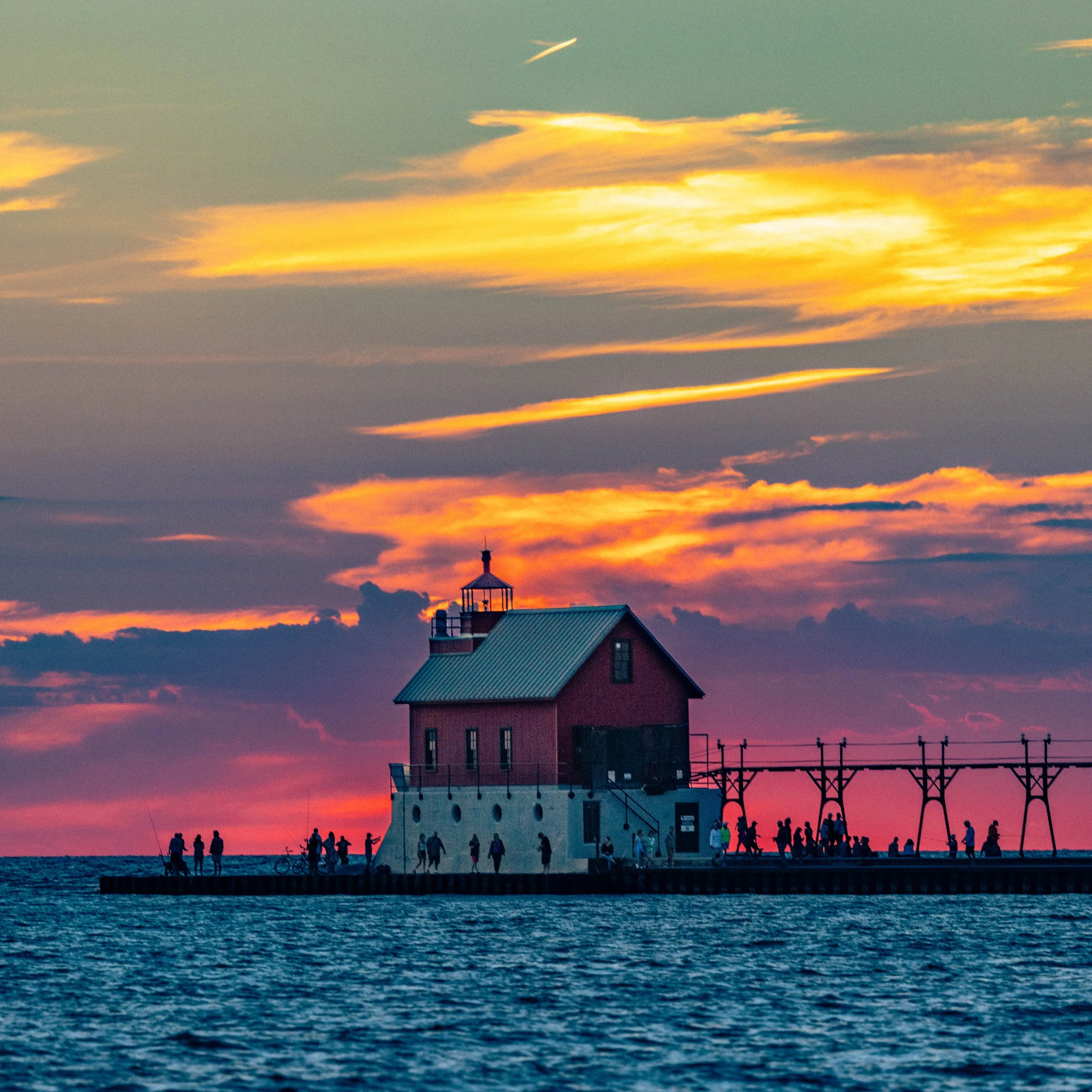 Sunset over the water with a lighthouse, people walking along the shoreline, and clouds illuminated in warm colors.