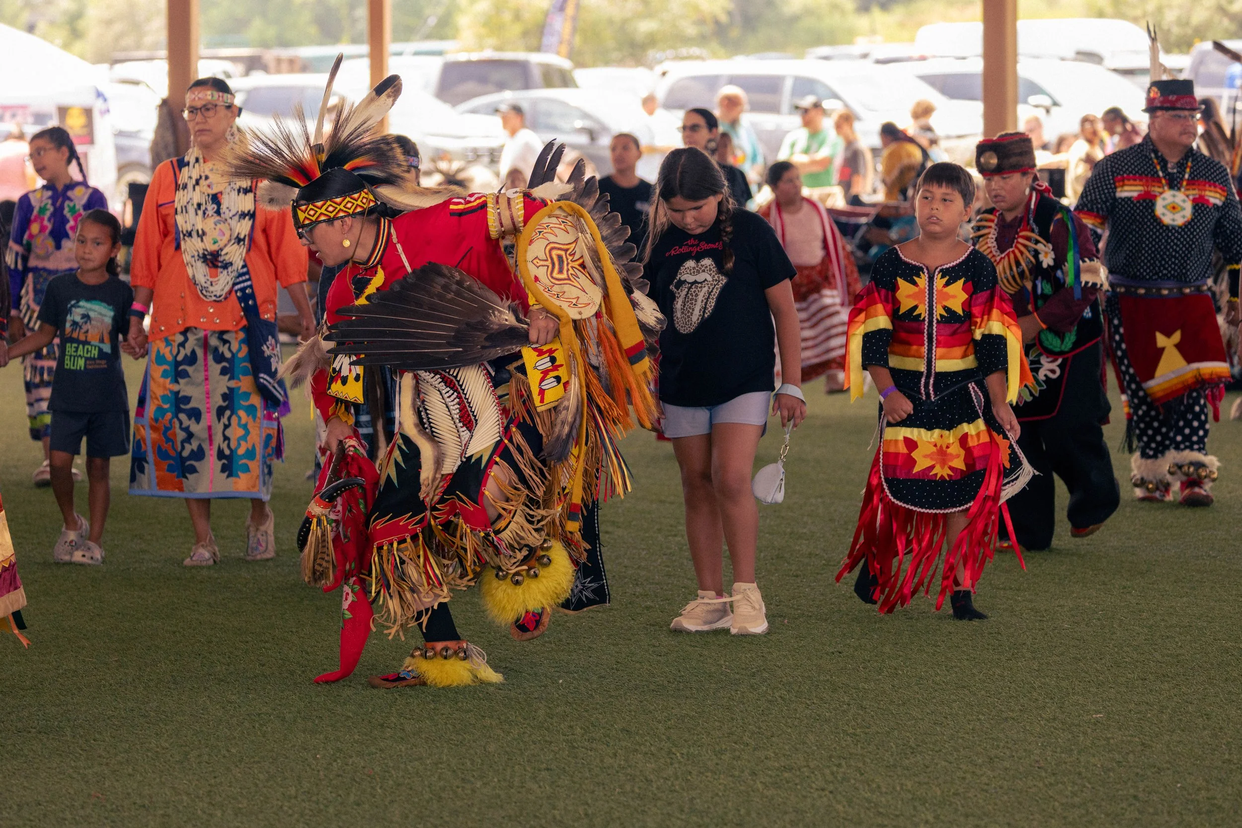 Native American dancers and participants at a cultural event, dressed in traditional regalia with feathered headdresses and colorful attire, under a pavilion with parked cars in the background.