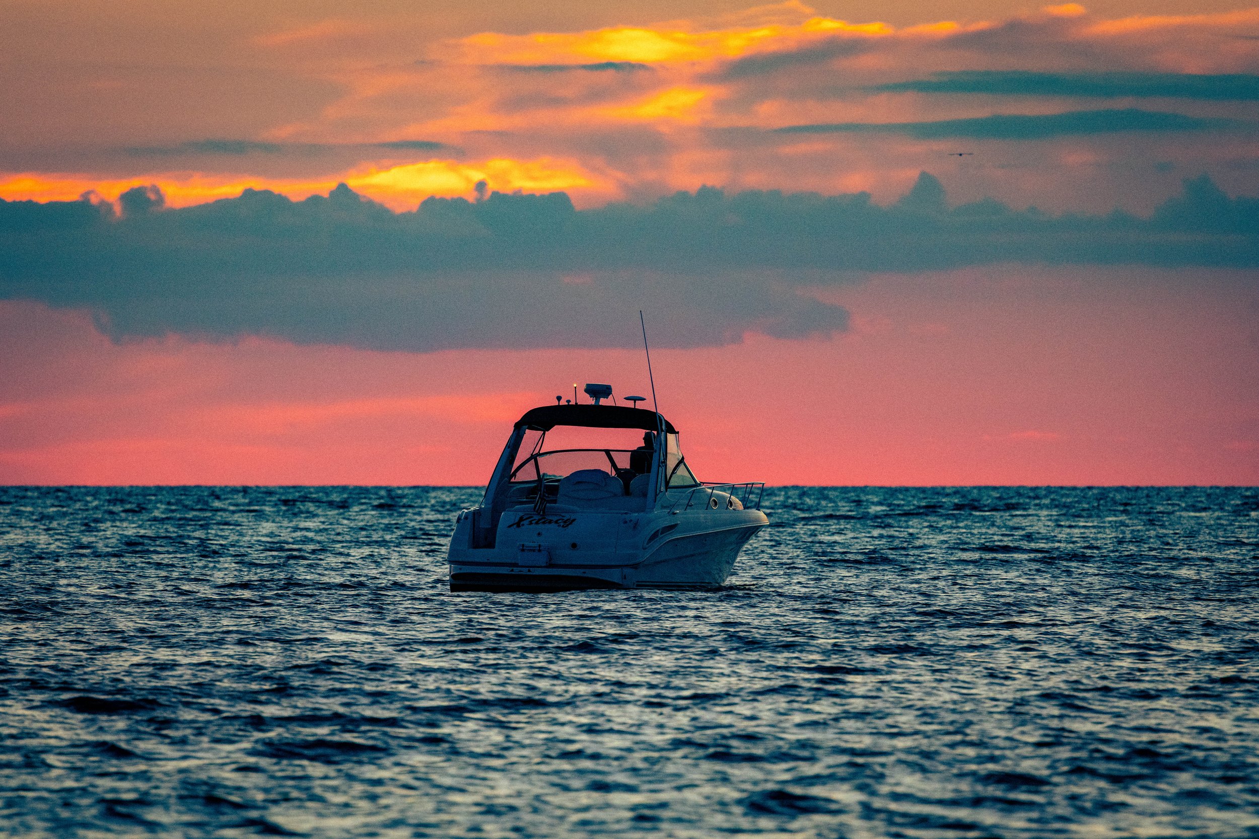 A boat floating on calm ocean waters during sunset with orange and pink clouds in the sky.
