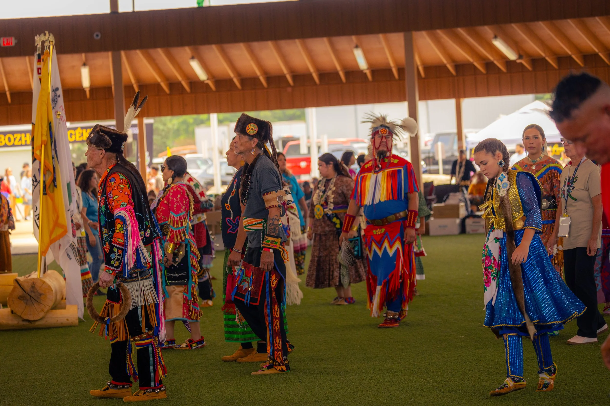A group of Native Americans dressed in traditional colorful regalia, participating in a cultural dance or ceremony inside a large wooden pavilion at an outdoor event.