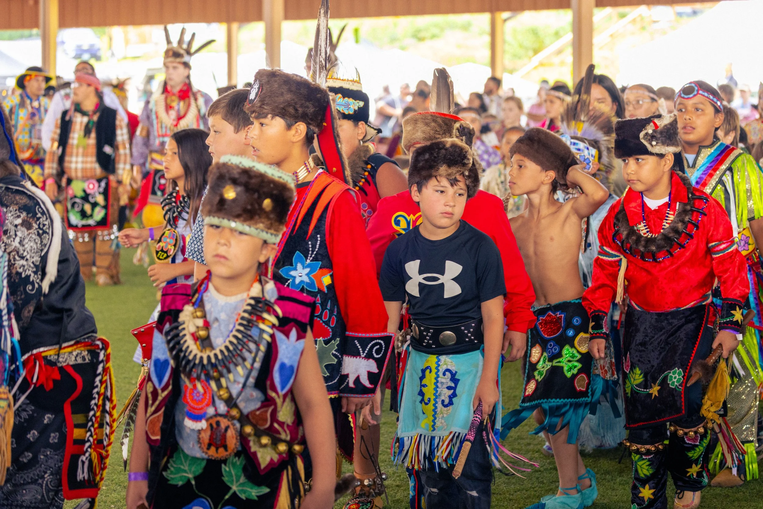 Children dressed in traditional Native American attire, standing in a line at a cultural event, with spectators in the background.