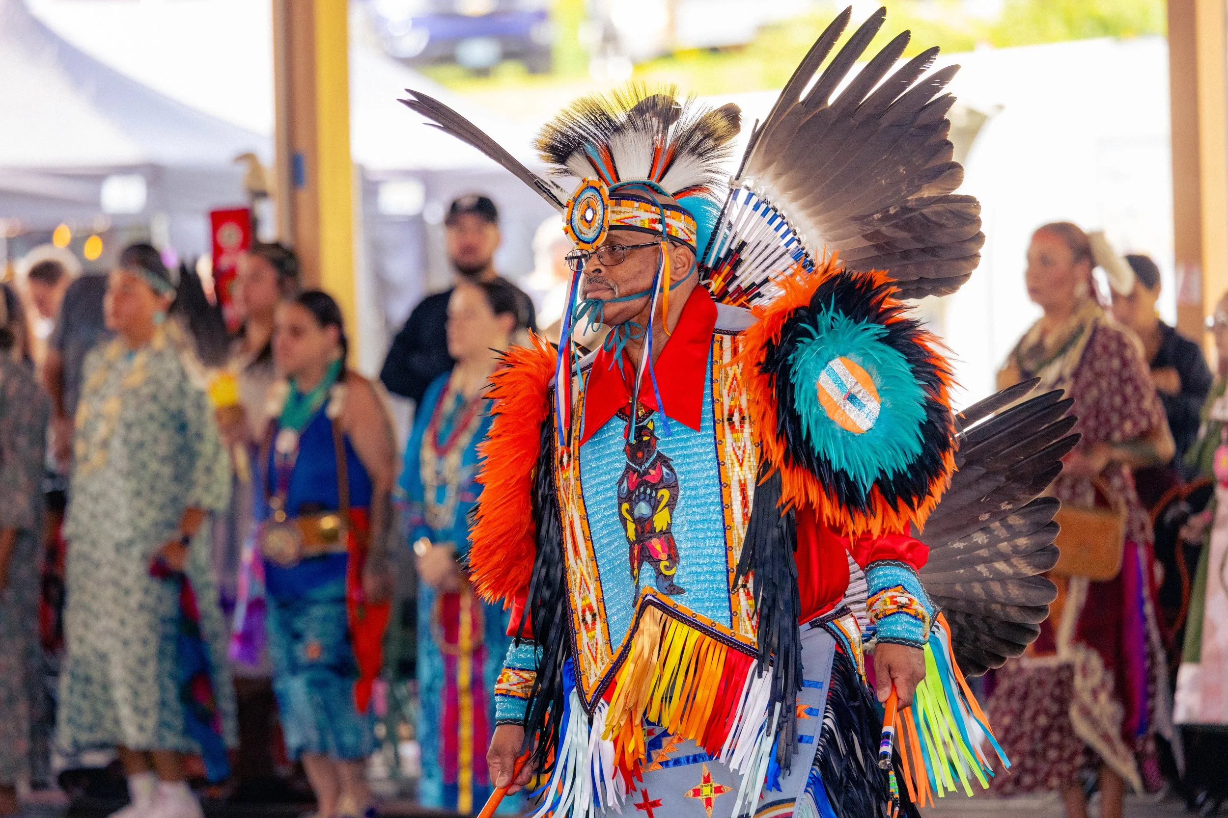 A Native American man in traditional feathered headdress and colorful clothing participating in a cultural celebration with a group of people in the background.