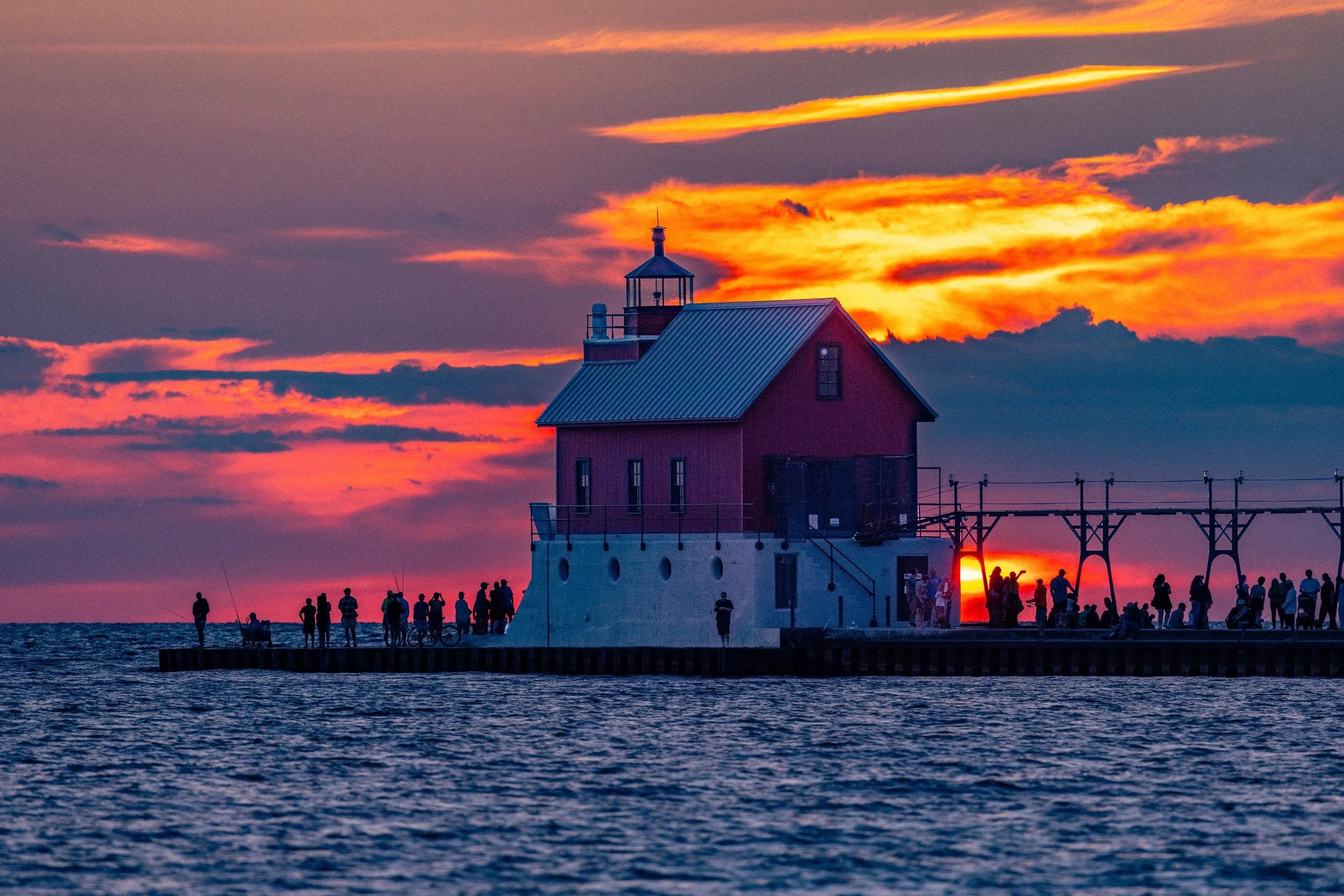Sunset over a lighthouse on a pier with silhouettes of people walking and fishing.