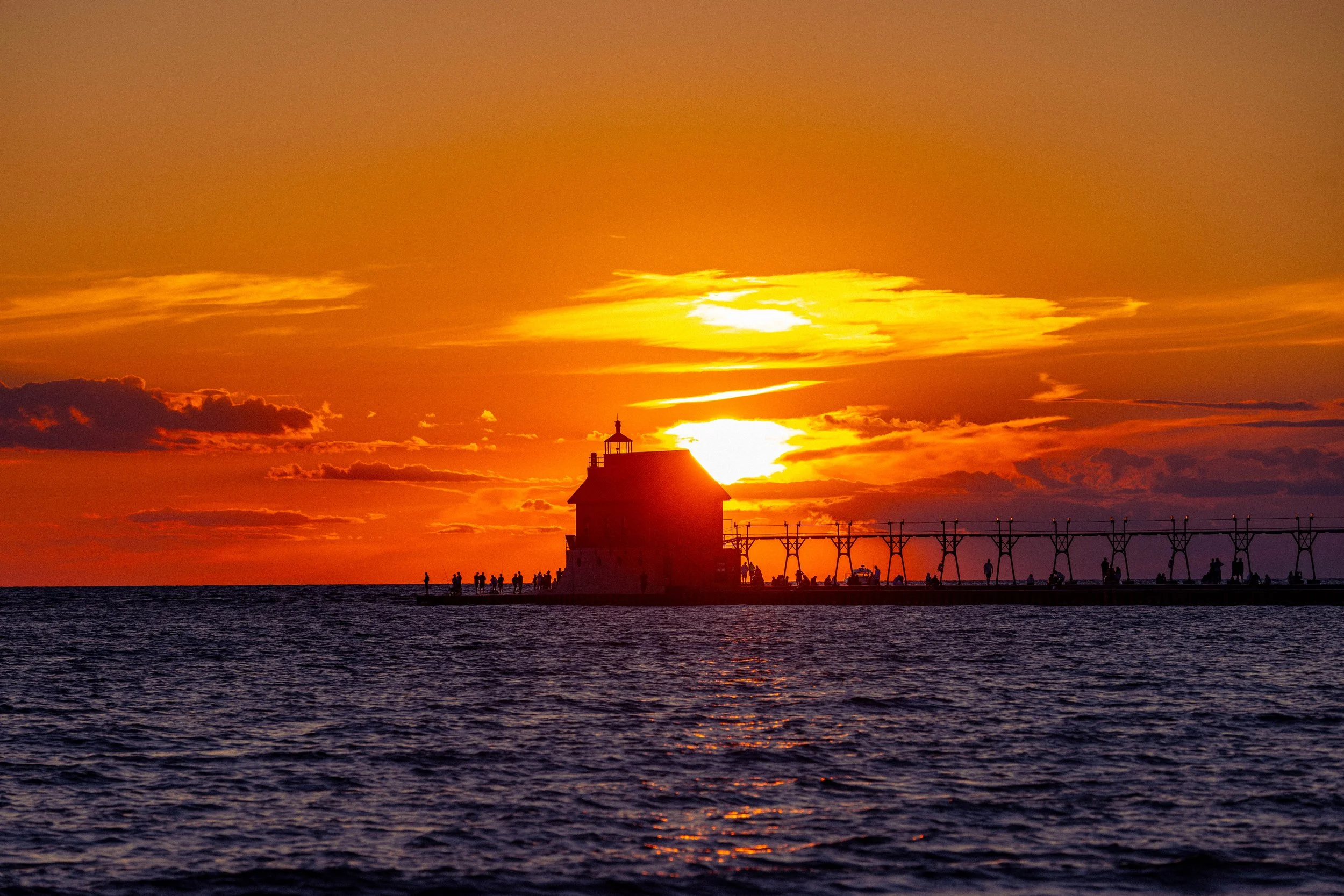 Sunset over water with dark clouds, small lighthouse, and people on a pier or breakwater in the foreground.