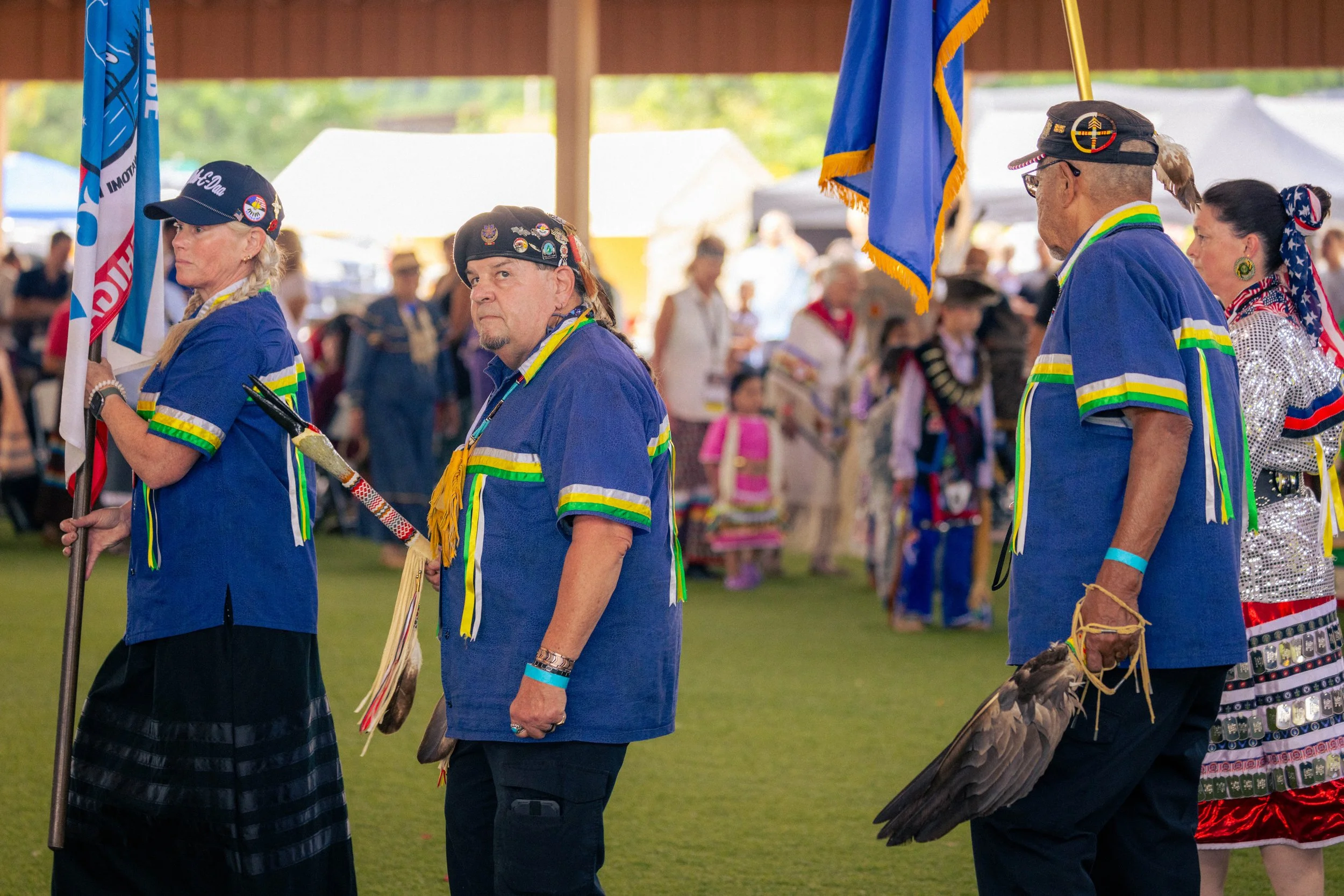 Group of Native American women and men dressed in traditional clothing, participating in a cultural dance or ceremony outdoors at a gathering or Pow Wow, holding flags and feathers.