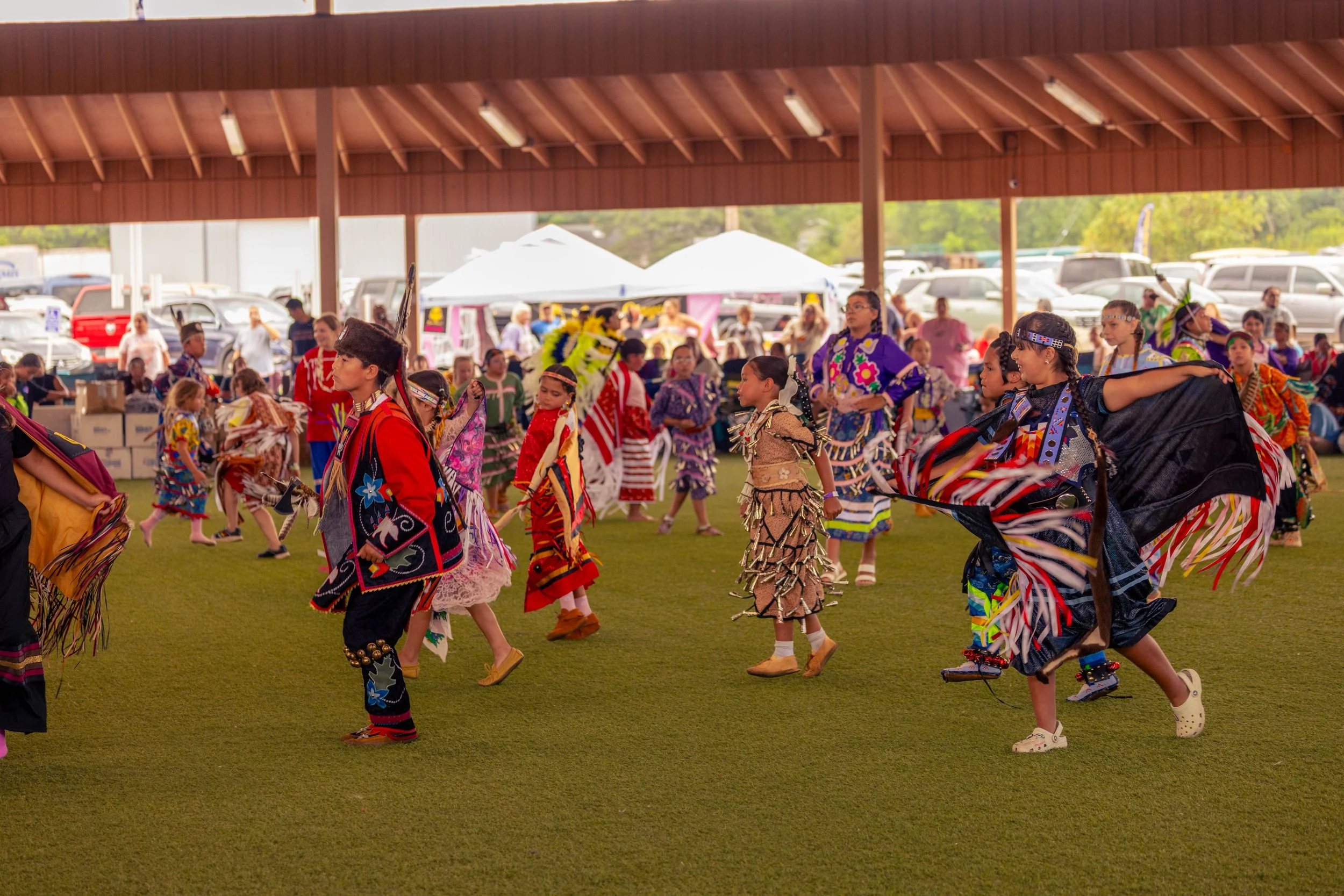 A group of children dressed in traditional Native American attire, dancing at an outdoor event under a large pavilion. There are tents, cars, and spectators in the background.
