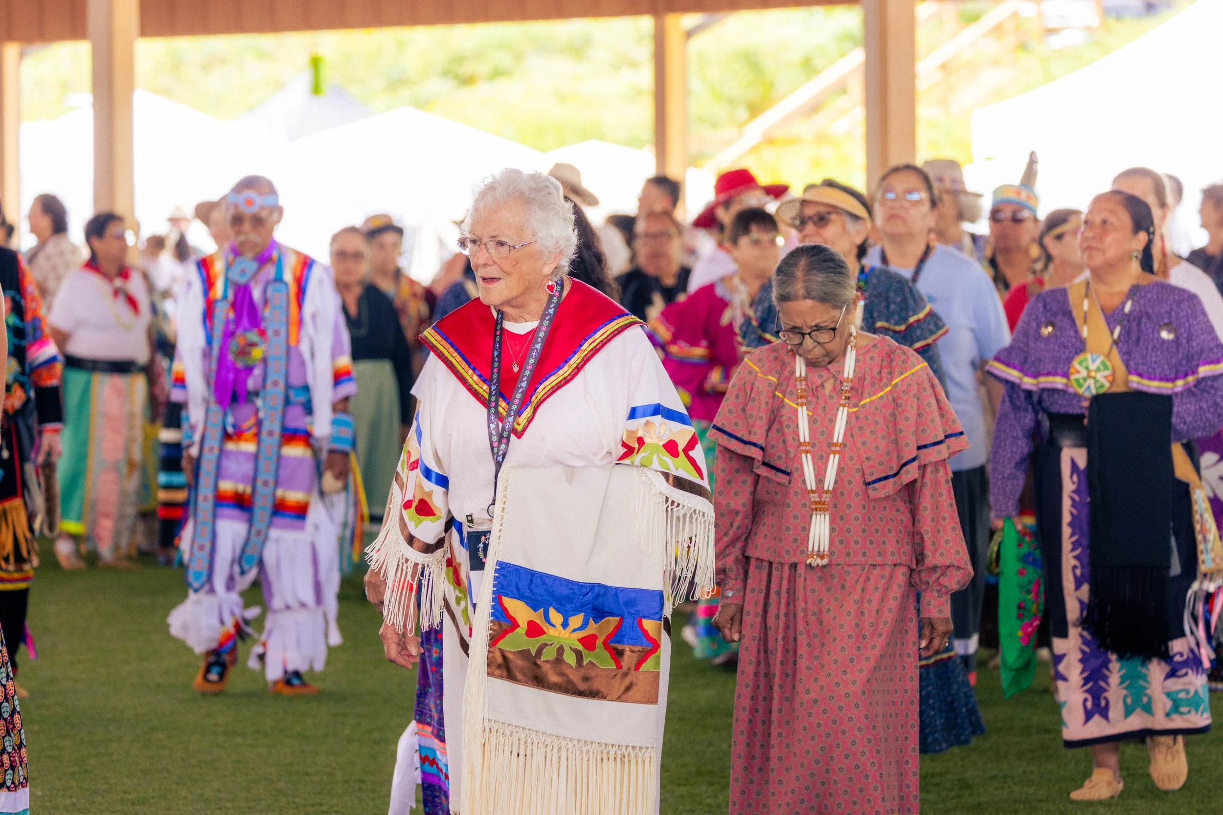 A group of Native Americans in traditional clothing participating in a cultural dance or ceremony inside a large wooden pavilion, with sunlight coming through the open sides.