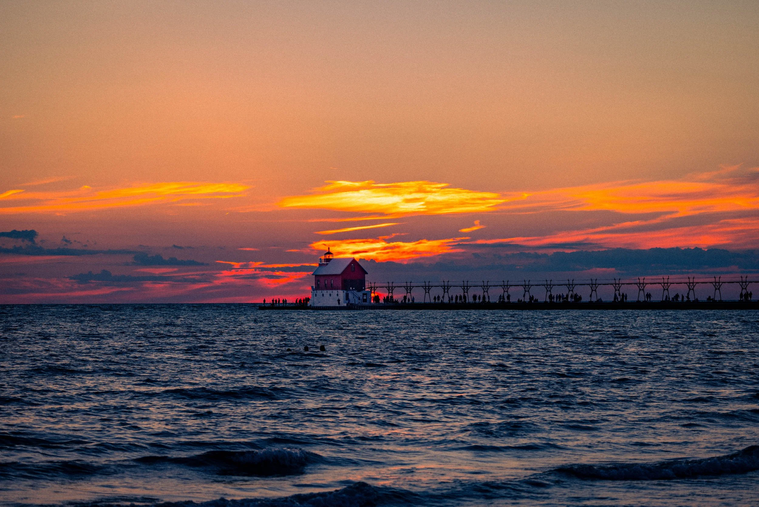 A sunset over the ocean with a lighthouse on a pier and a bridge, and a group of people walking along the pier and the shoreline.