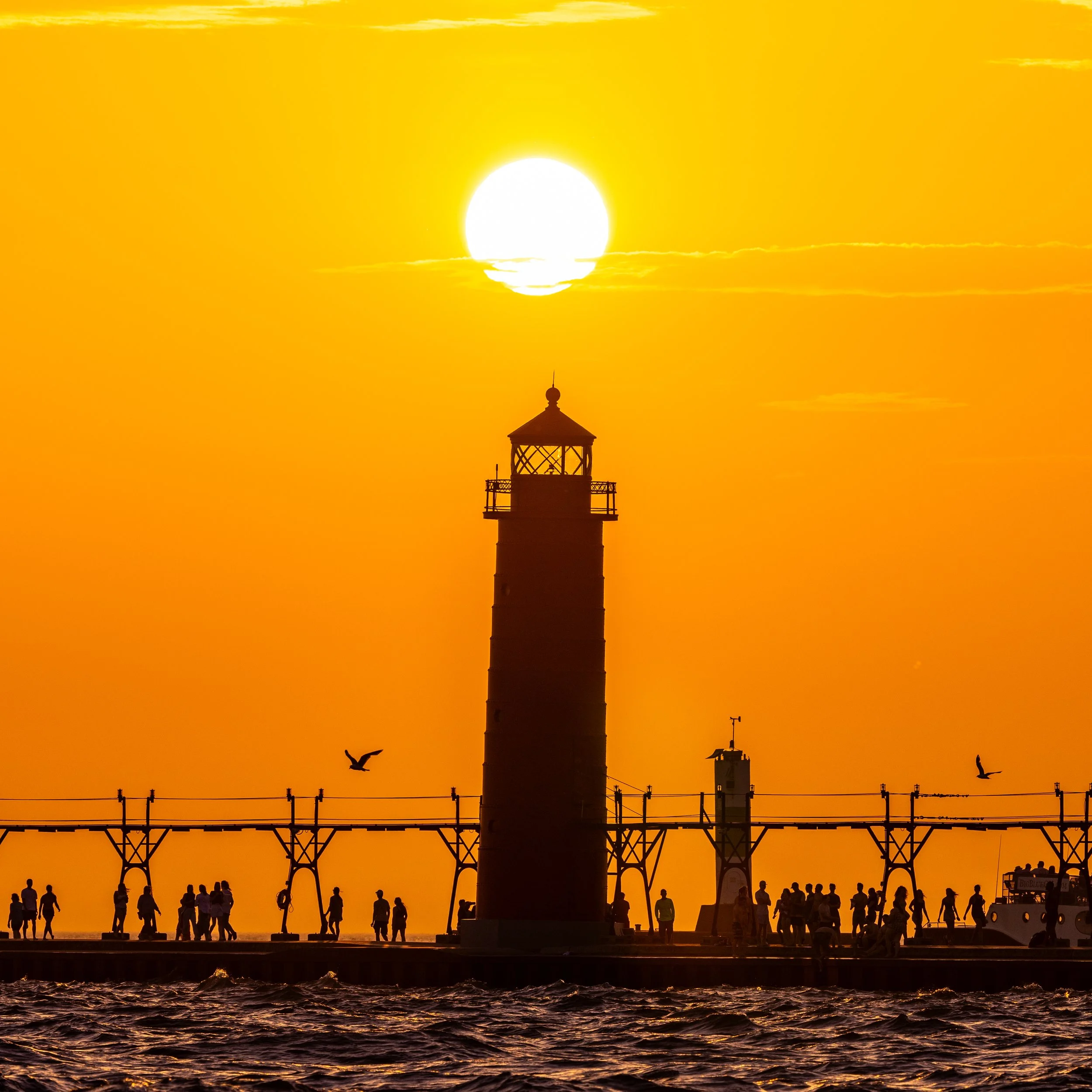 Sunset over a pier with a lighthouse and people walking along the water, birds flying in the sky.