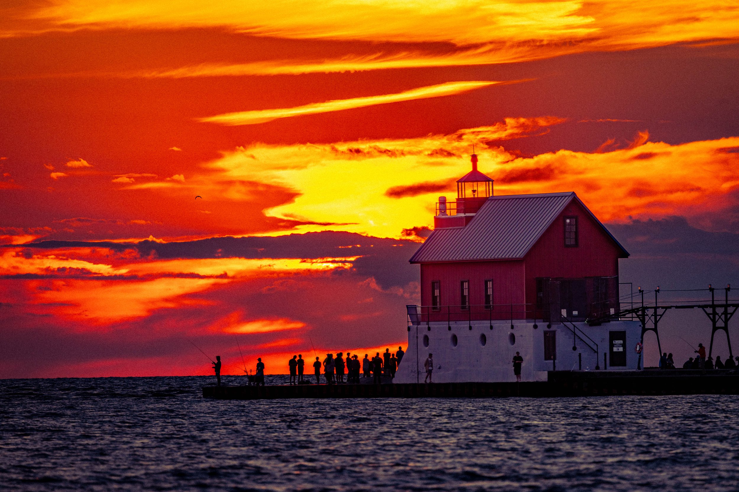 A lighthouse by the water during a vibrant sunset with orange and yellow clouds. Silhouettes of people are standing on a pier, some fishing, with a bird flying nearby.