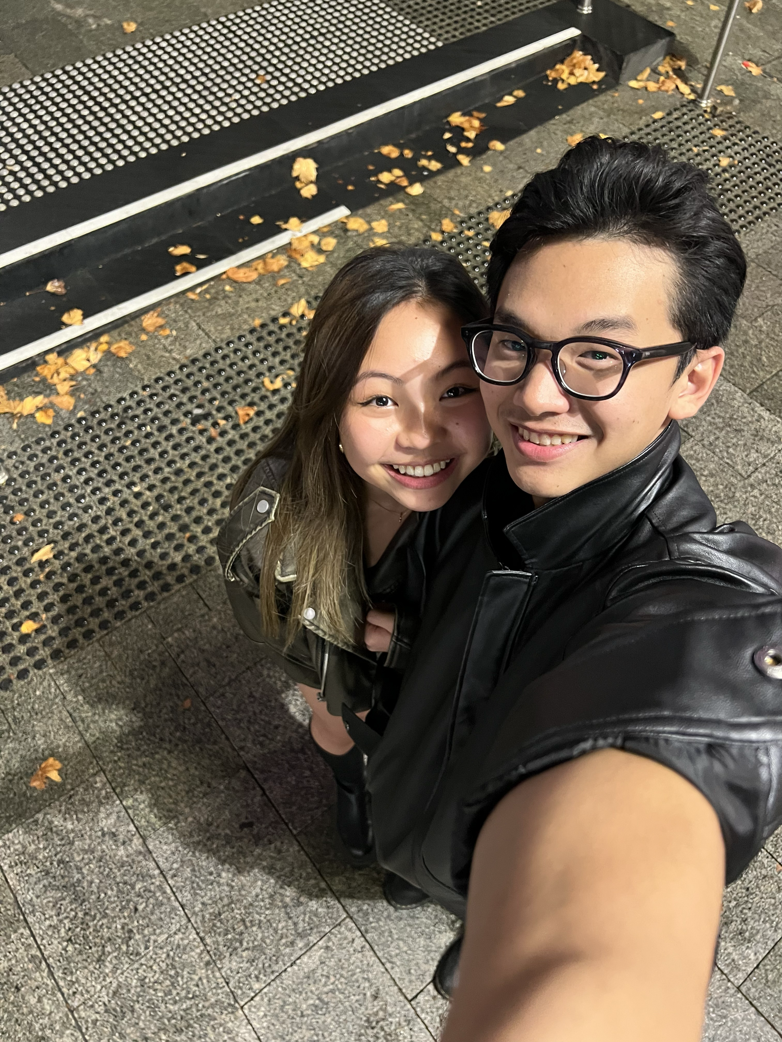A selfie of a smiling young woman and a young man with glasses, both dressed in black leather jackets, standing on a sidewalk with fallen autumn leaves. The photo is taken from a high angle with the person's arm visible in the foreground.