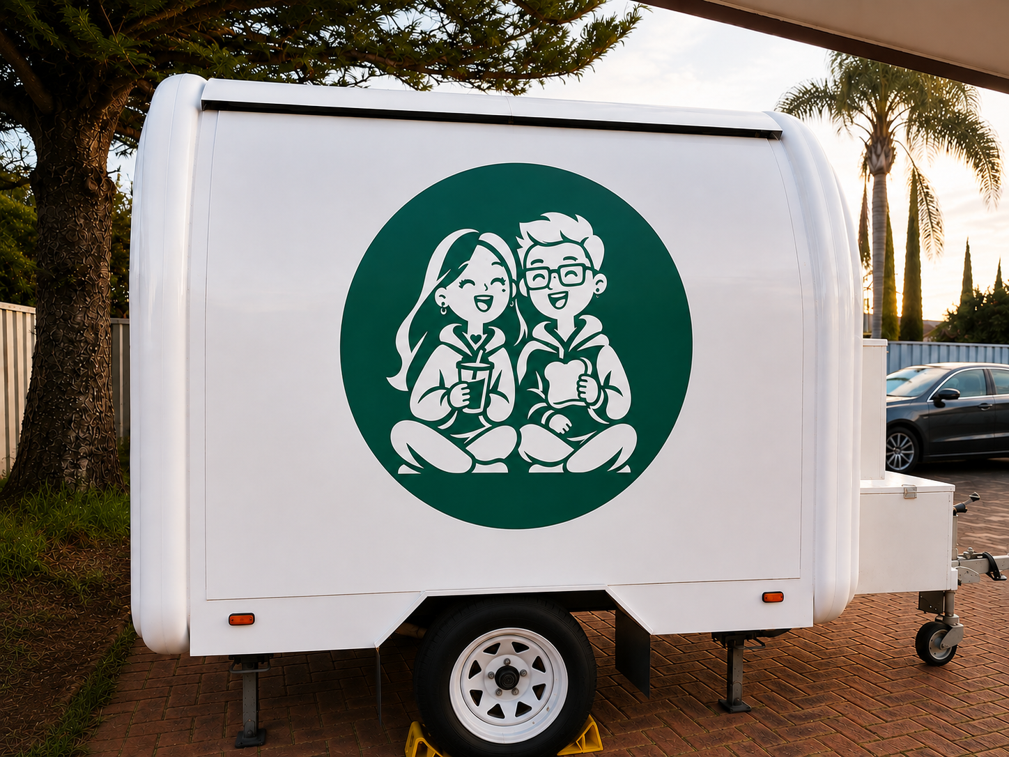 A food truck with a logo depicting a cartoon girl and boy sitting cross-legged, smiling, and holding food and a drink, set outdoors with trees and parked cars in the background at sunset.