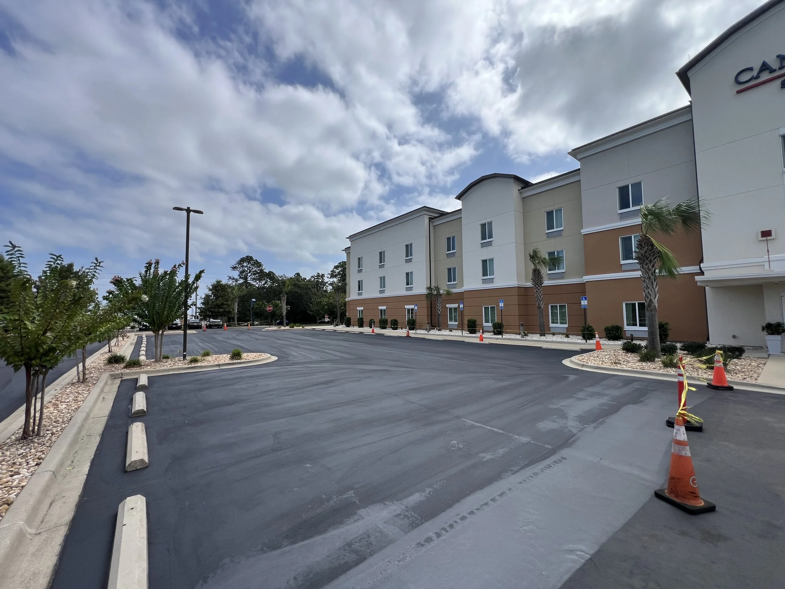 A parking lot outside a multi-story building with a partly cloudy sky, scattered trees, and orange traffic cones.