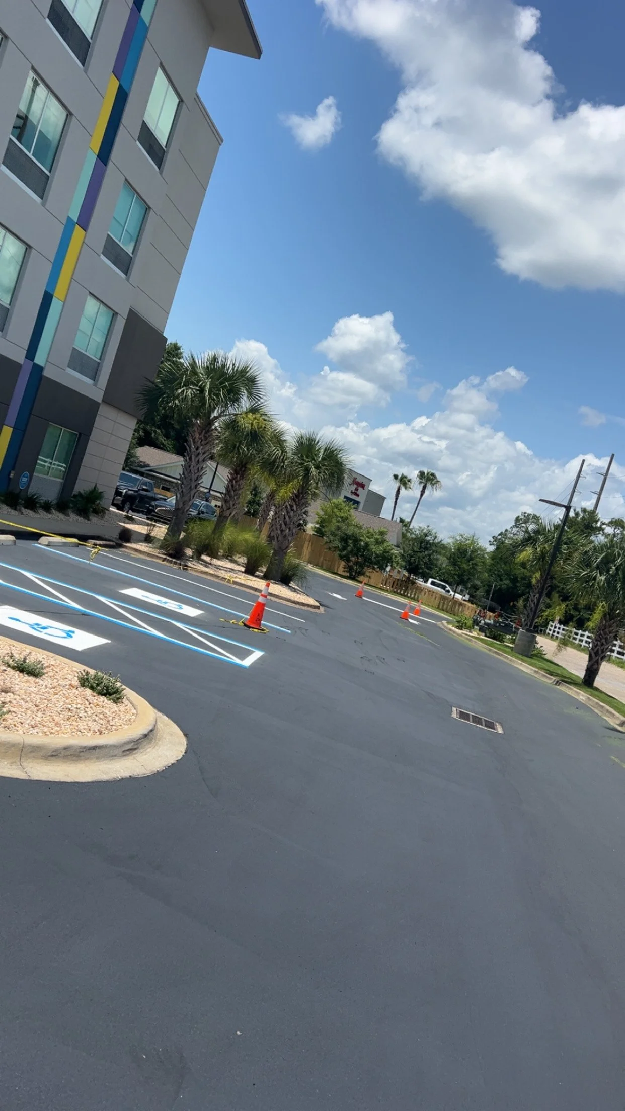 A parking lot with newly painted handicapped parking spaces marked in blue and white. The lot has orange cones set up around the spaces, and there are palm trees, a multi-story building with a colorful stripe design on the side, and a partly cloudy sky in the background.