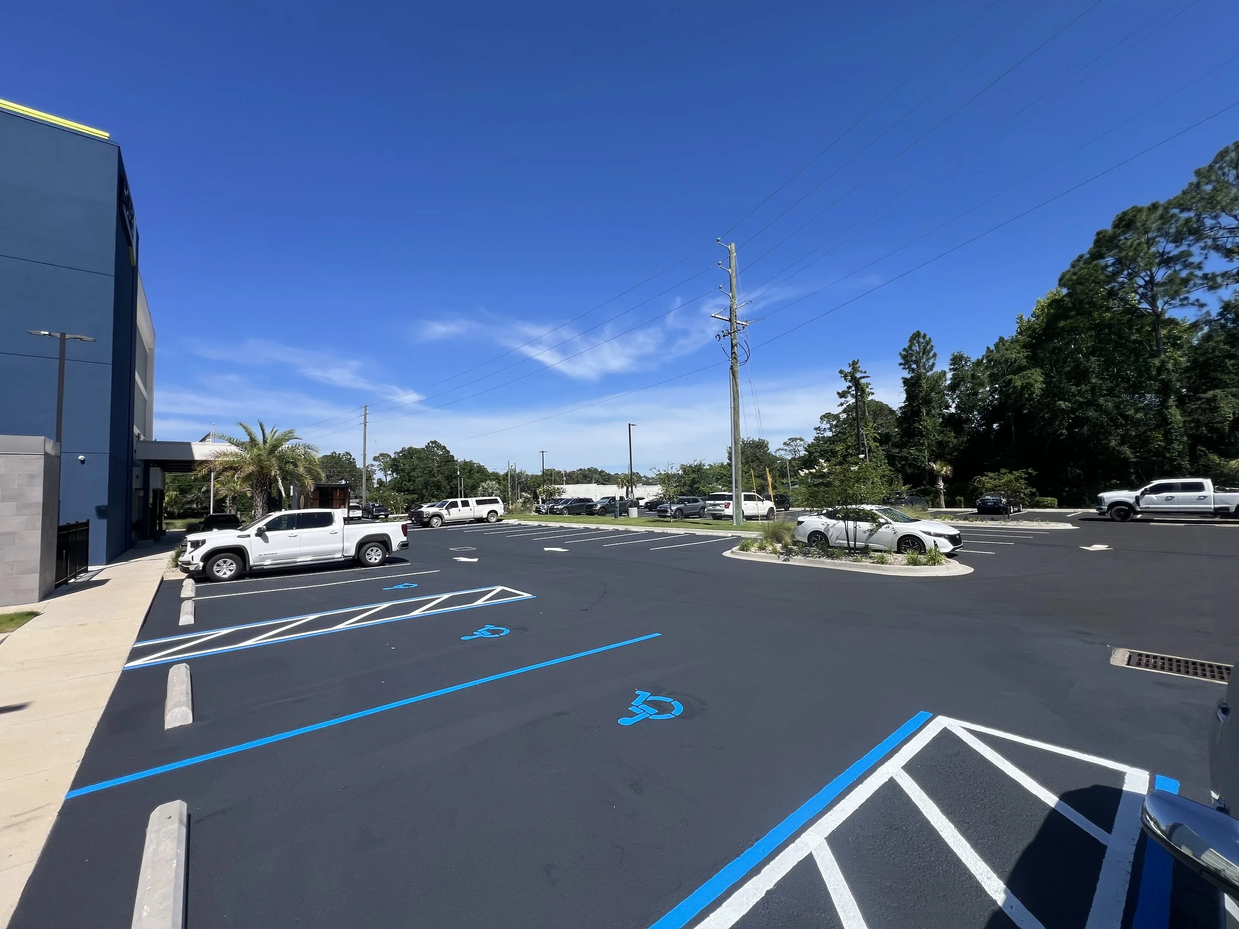 A parking lot with several empty handicapped parking spaces marked with blue paint and wheelchair symbols. Cars are parked in the background, and the lot is bordered by trees and a commercial building on the left. The sky is clear and blue.