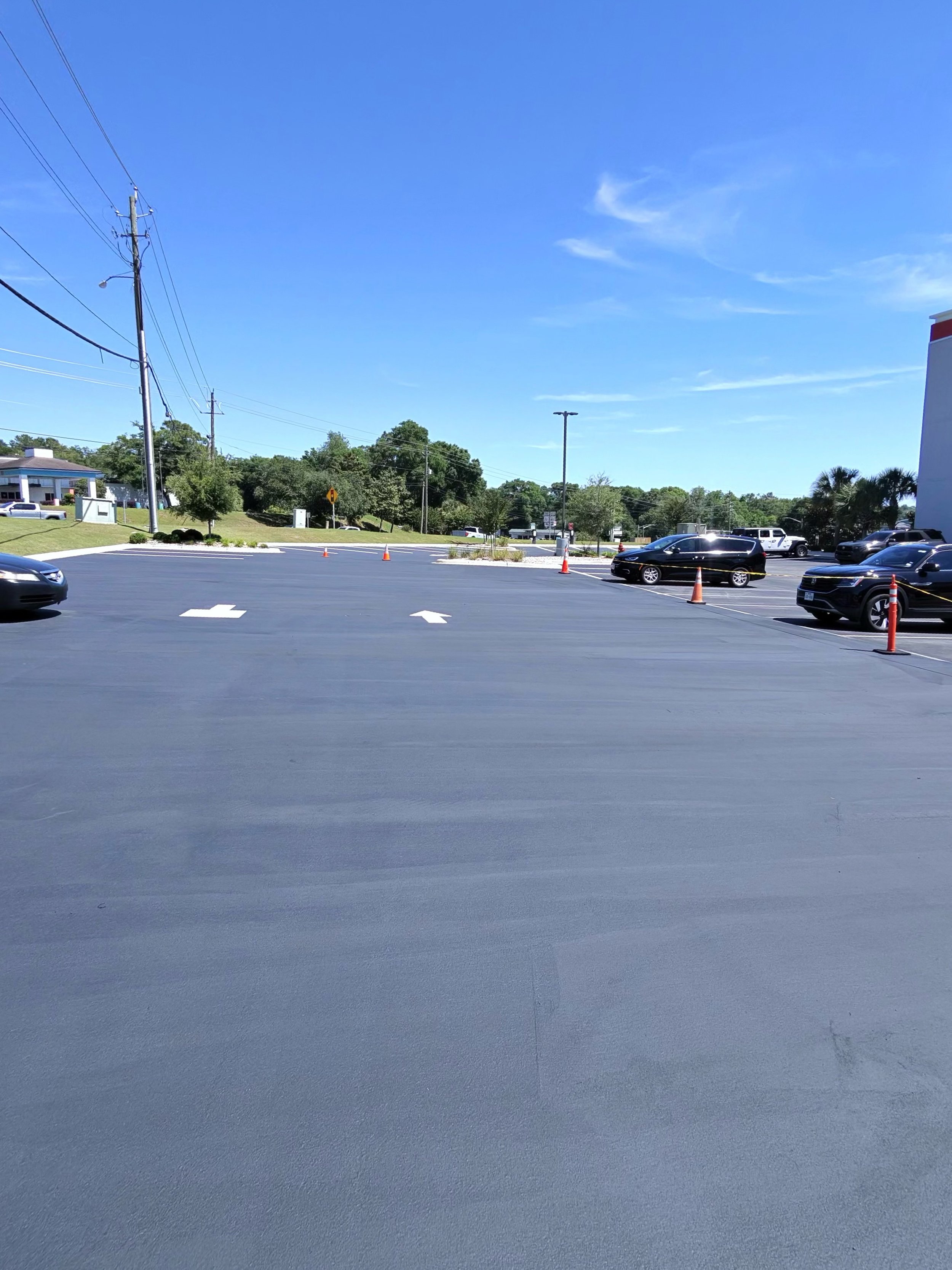 A parking lot with several cars and orange traffic cones, trees, and a building under a clear blue sky.
