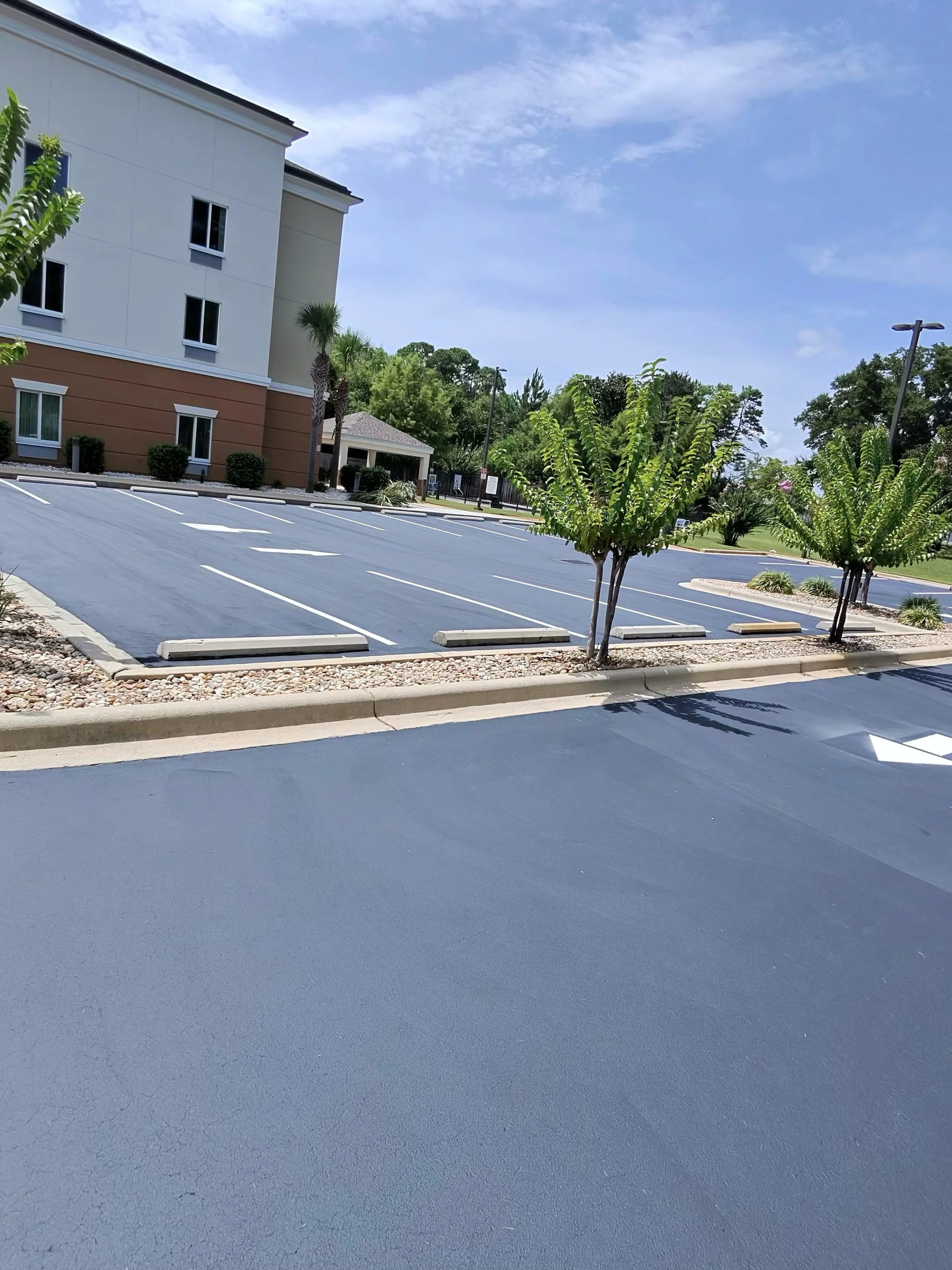 Empty parking lot with newly paved asphalt, trees, and a building in the background under a partly cloudy sky.