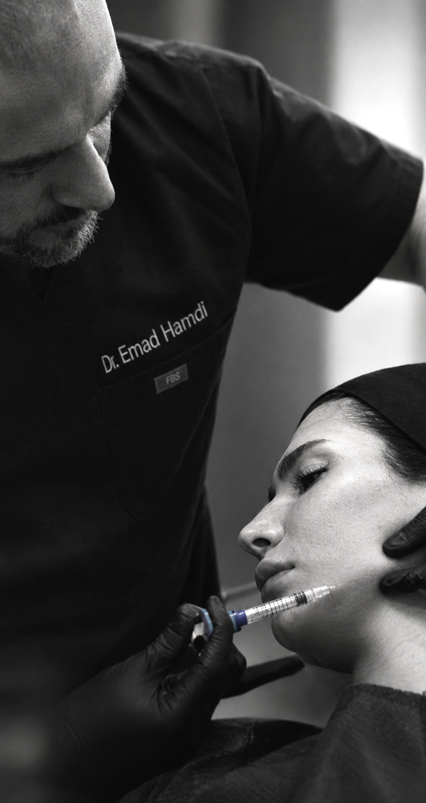 A medical professional administering a cosmetic injection to a woman's face during a procedure, with the professional wearing a uniform labeled with "Dr. Emad Hamdi".