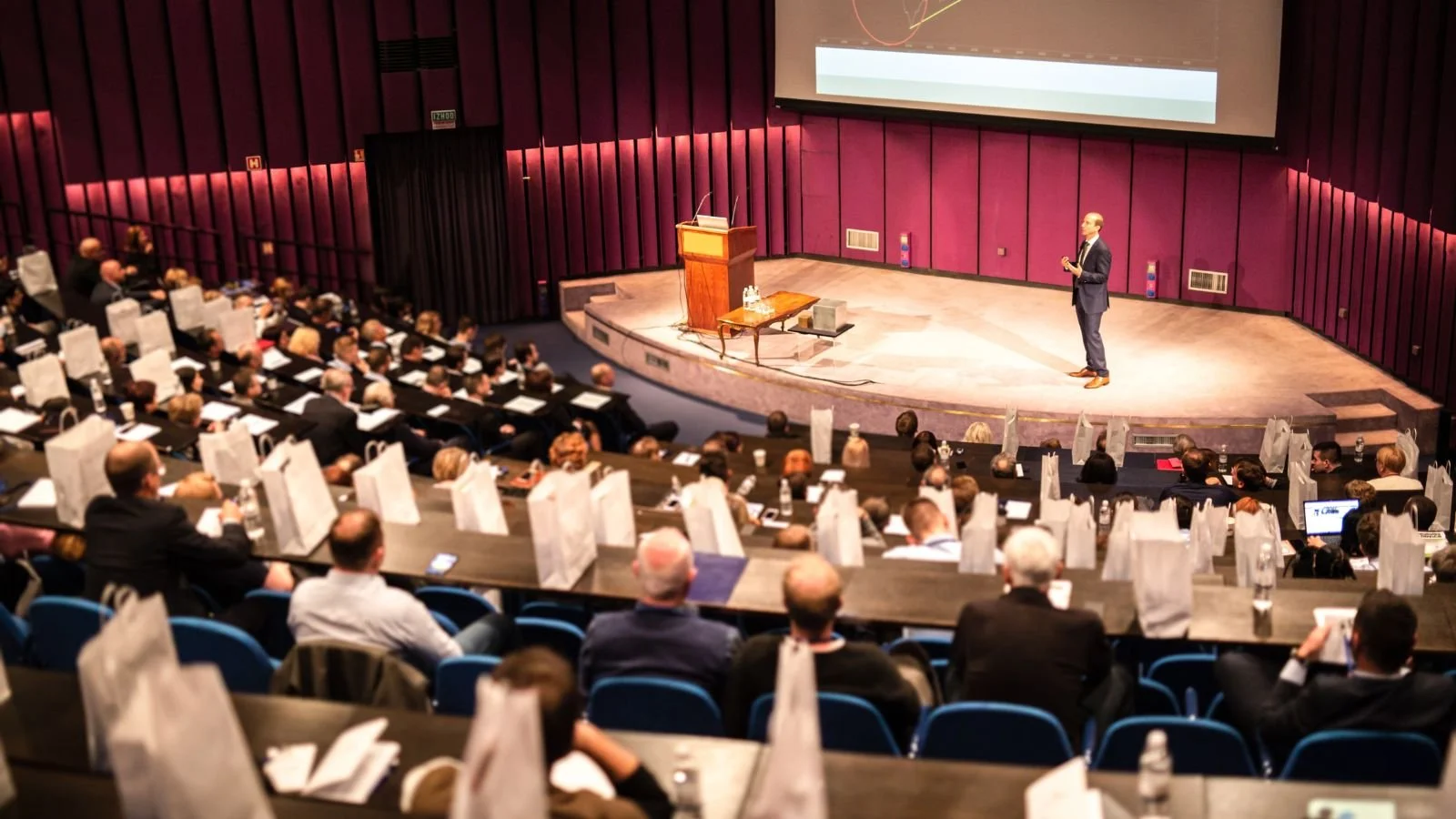A man in a dark suit giving a presentation on stage in a large auditorium filled with seated audience members. The stage has a podium, a small table with bottles of water, and a large screen behind the speaker. Audience members are seated at long tables with white gift bags, bottles of water, and notebooks.
