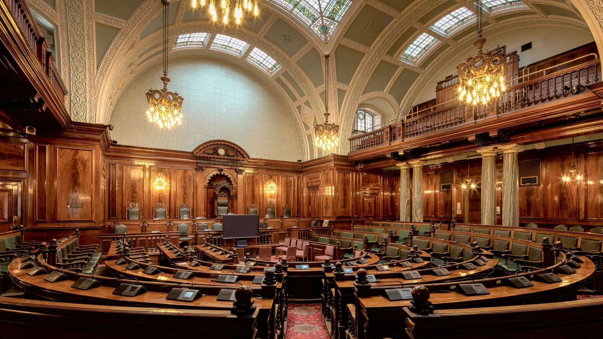 Interior of a grand courtroom with high vaulted ceilings, arched windows with stained glass, ornate chandeliers, wood-paneled walls, and rows of green seats with microphones in front of them.