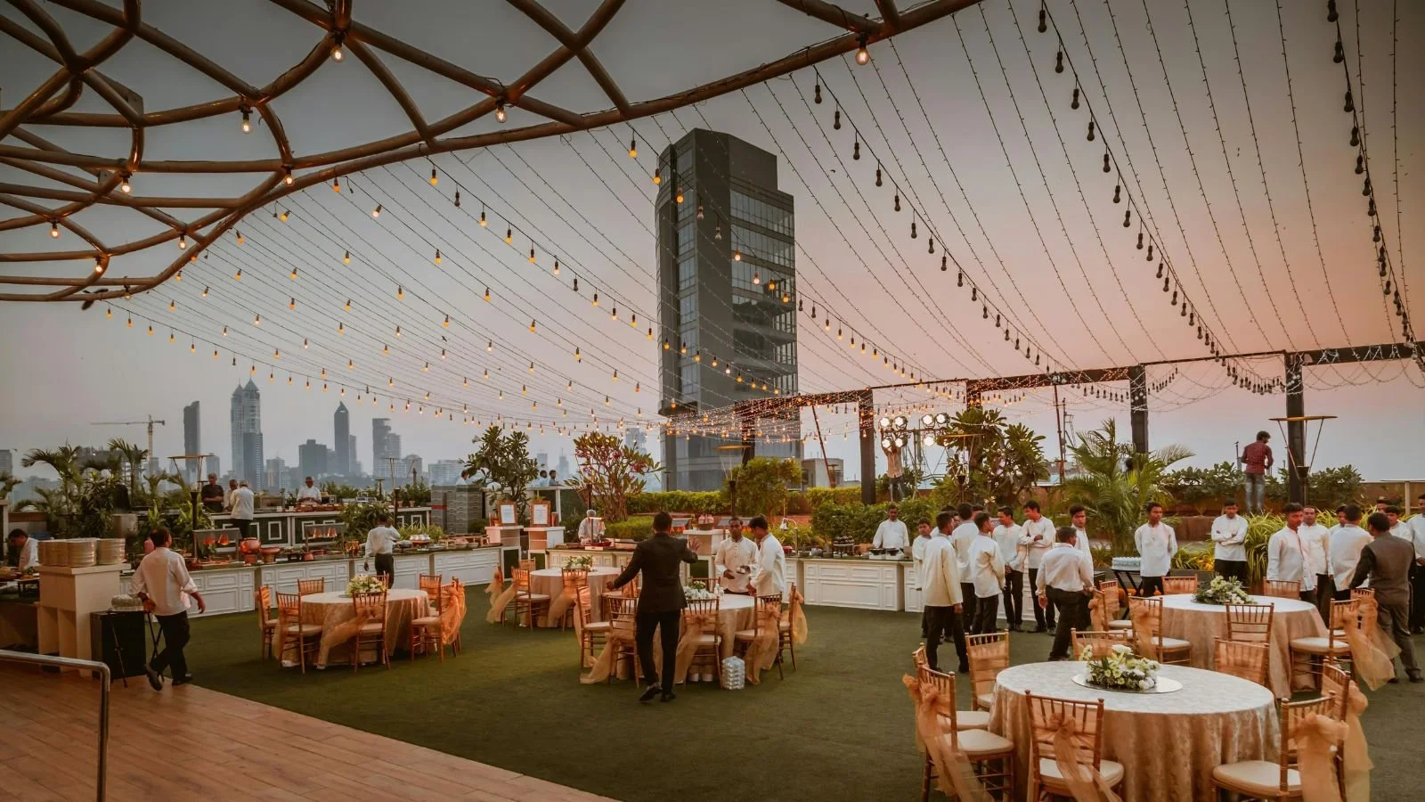 An outdoor rooftop event space with string lights and a city skyline in the background during sunset, featuring round tables with gold-colored tablecloths and chairs, and people dressed in formal attire.