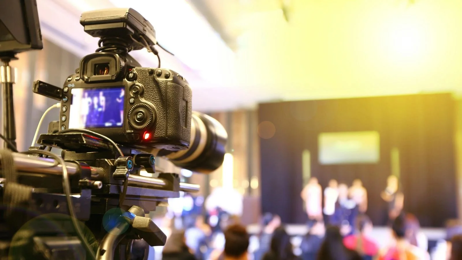 Camera on a tripod recording a stage with people standing and a large screen in the background at an event.