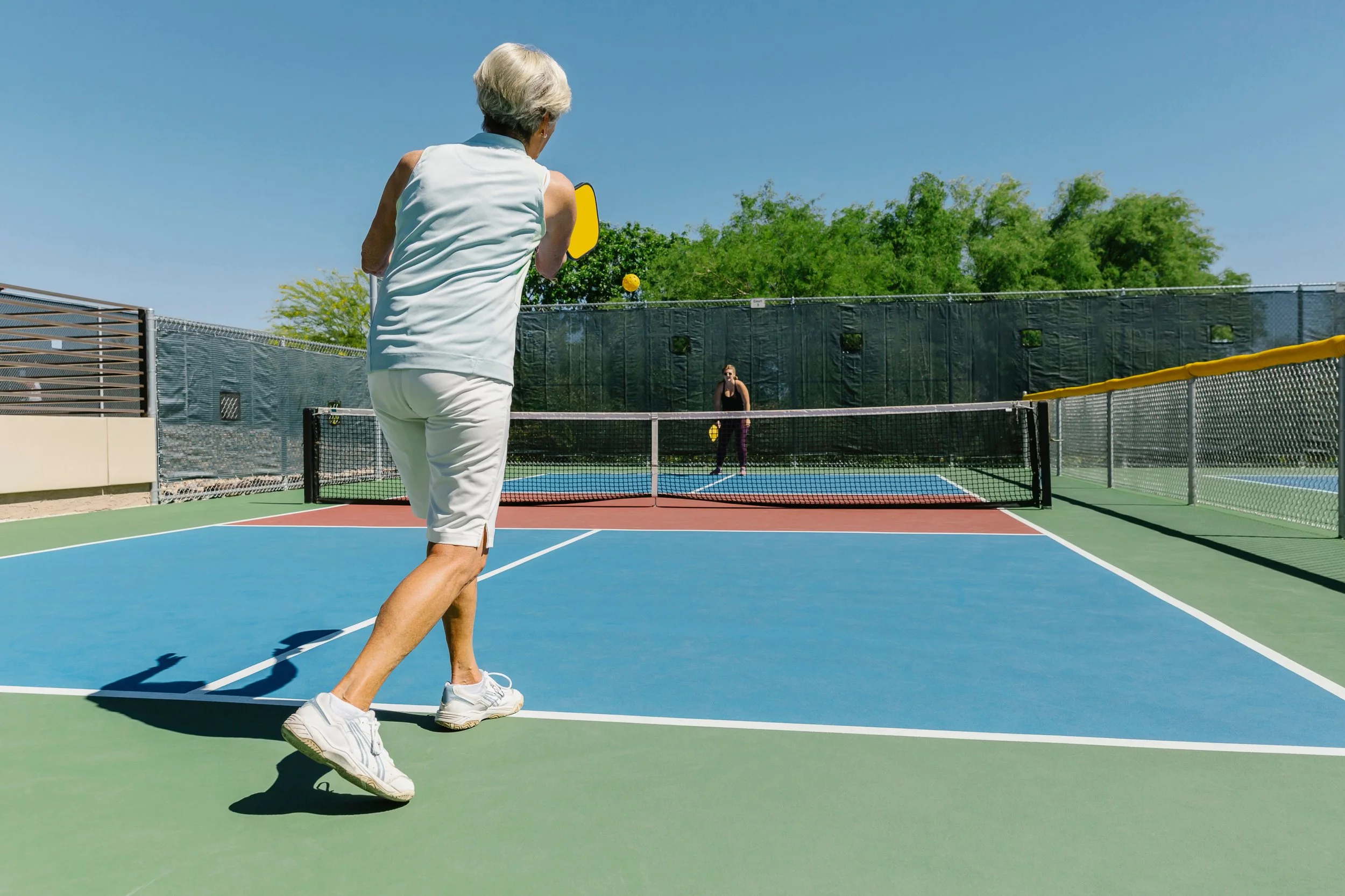 An elderly woman playing pickleball on an outdoor court, hitting the yellow ball with a paddle. Another person is visible on the opposite side of the net, and the court is surrounded by green trees under a clear blue sky.