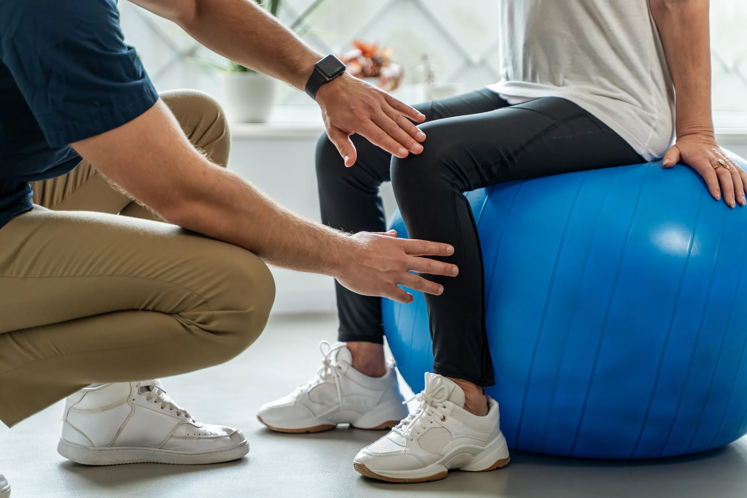 A physical therapist or trainer assisting a person seated on an exercise ball, positioning their leg for physical therapy or stretching exercise, in a well-lit indoor setting.
