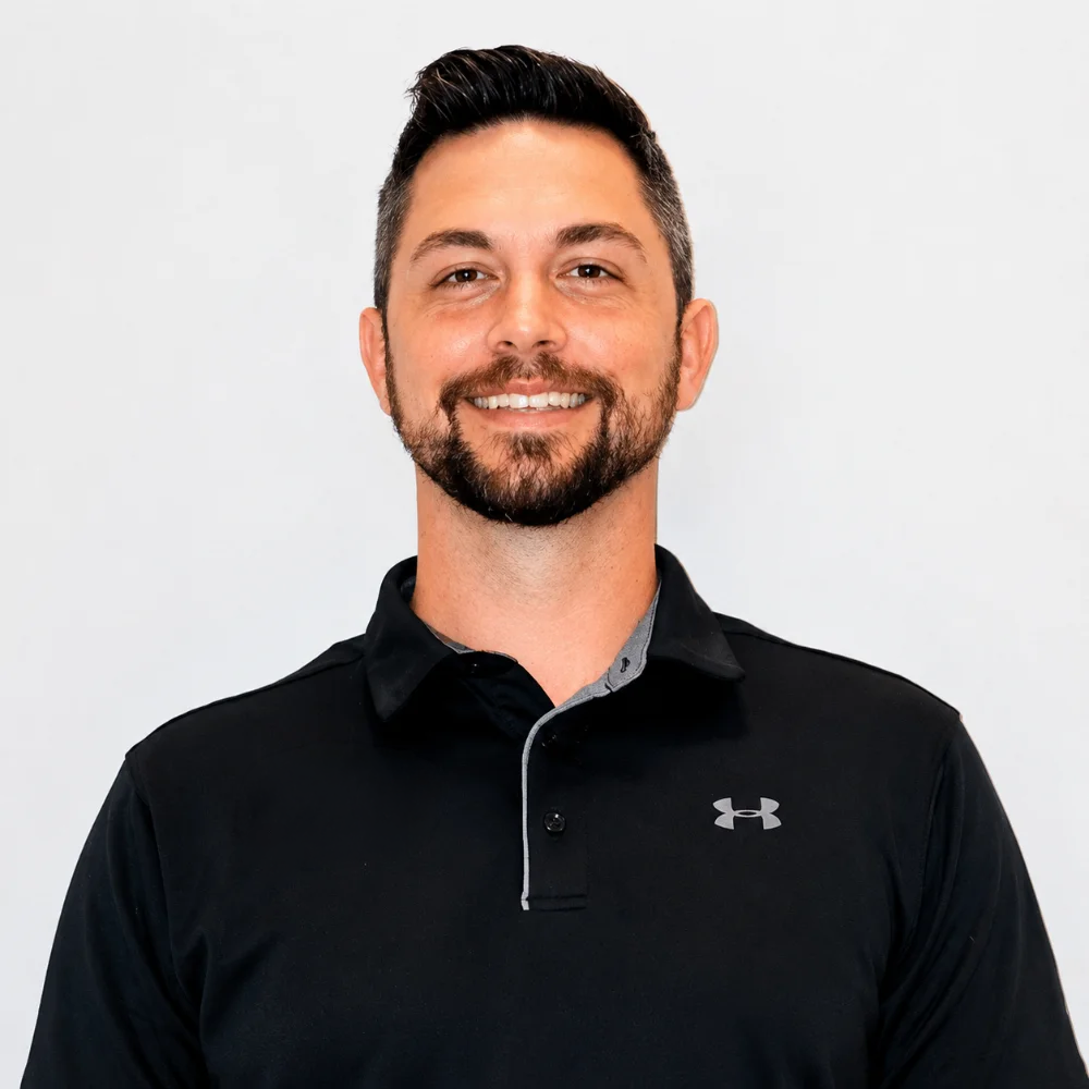 A man with short dark hair, a beard, and a mustache, smiling, wearing a black Under Armour polo shirt, standing against a plain white background.