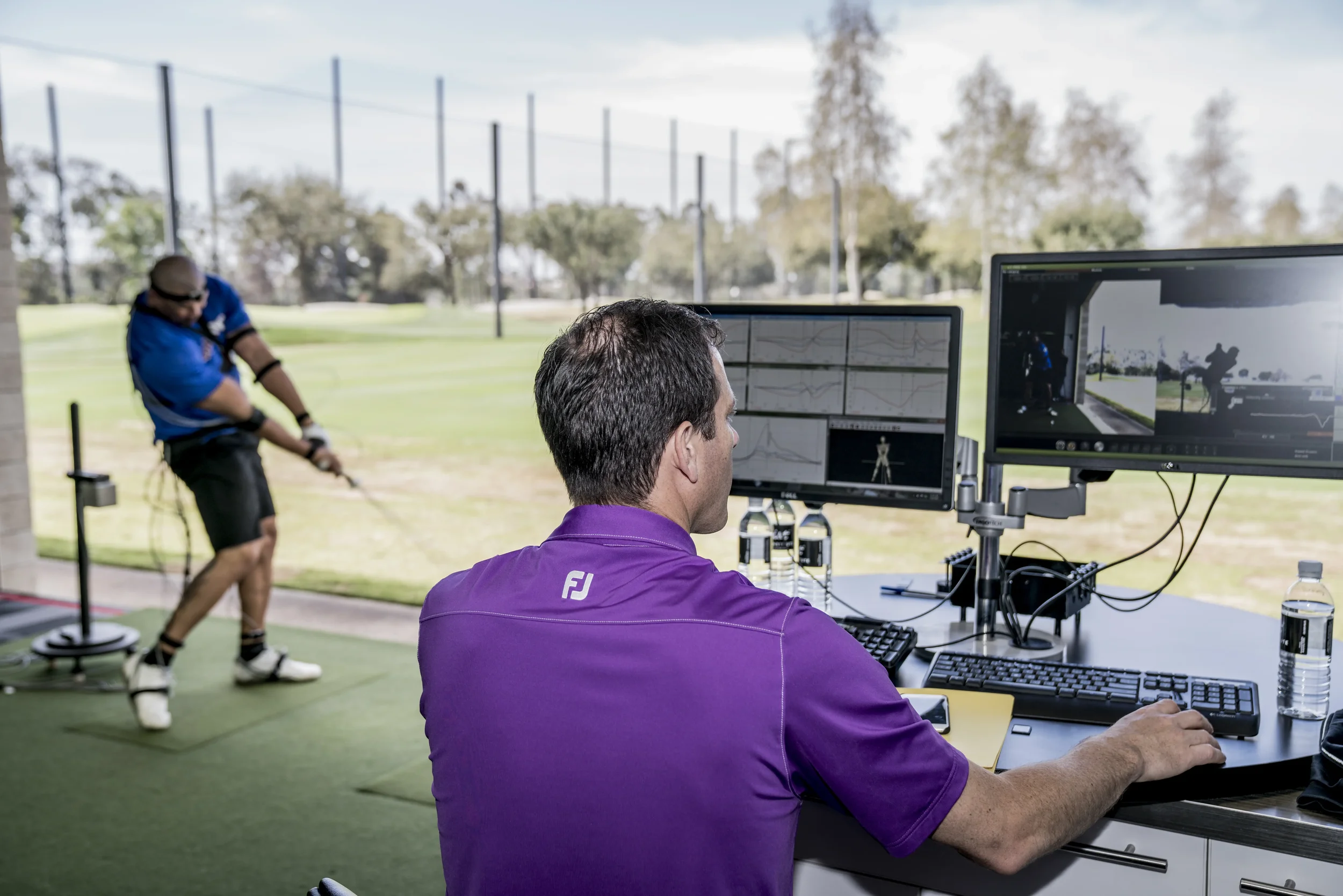 A man in a purple shirt working at a desk with multiple monitors, while a golfer in a blue shirt and black shorts hits a golf ball on an outdoor driving range in the background with trees under a cloudy sky.