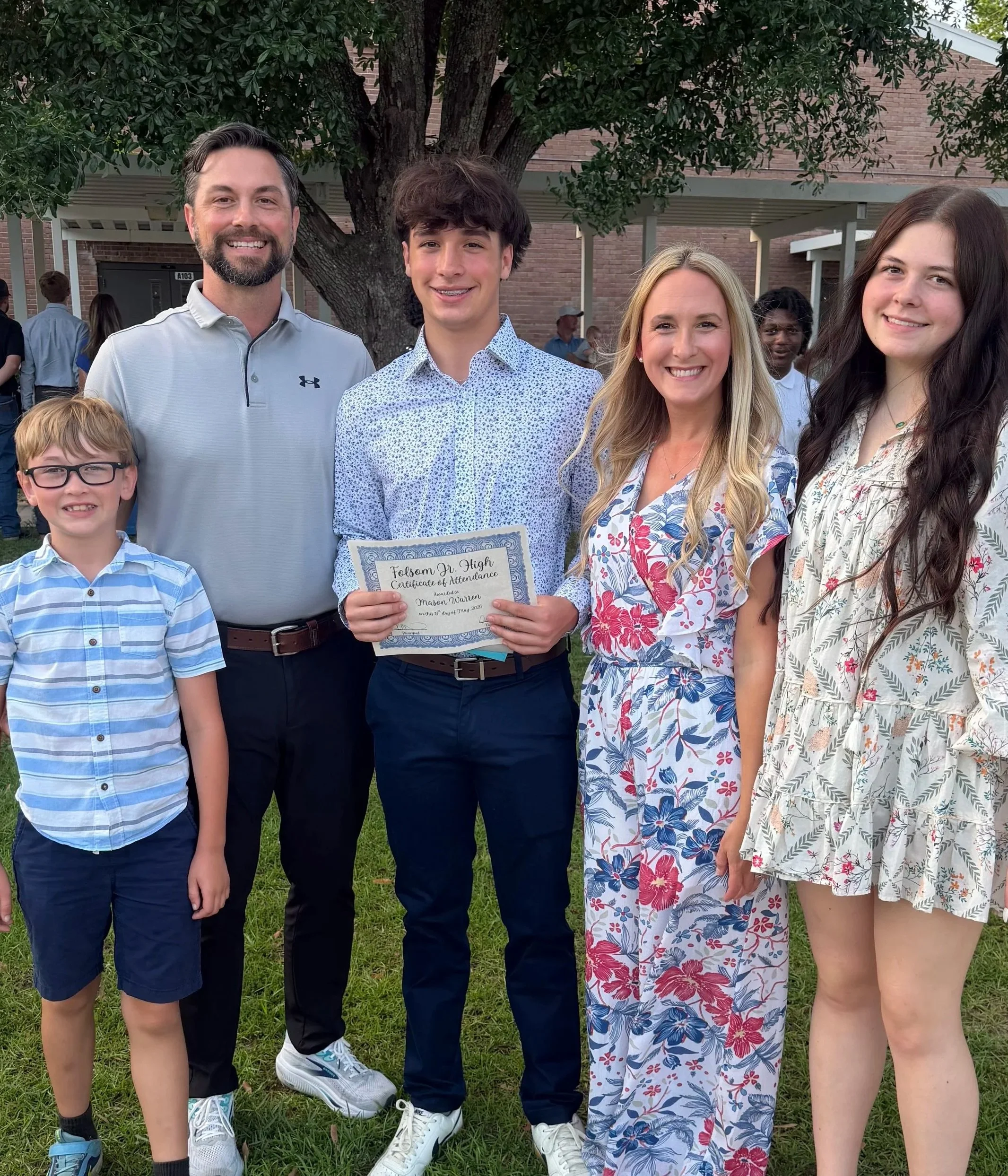 A group of four people, including a young man holding a certificate, standing outdoors under a tree with green foliage. They are smiling and dressed in casual summer clothing.
