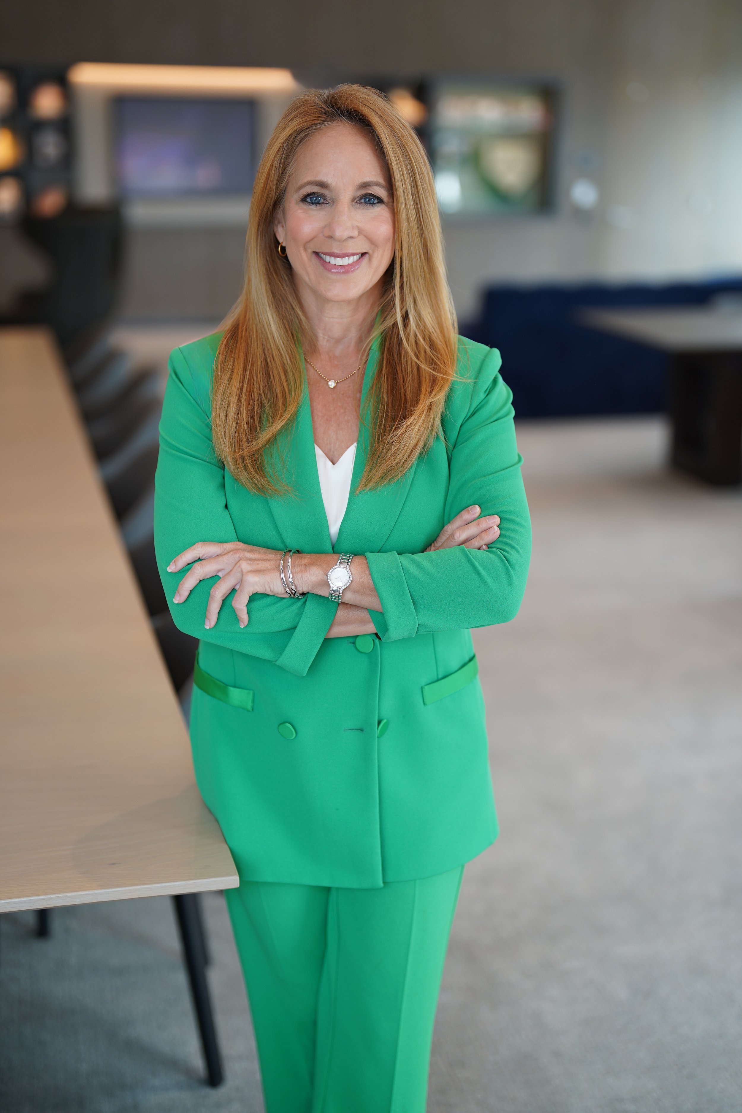 A woman with long red hair, wearing a green suit, is standing in an office. She is smiling, with her arms crossed, and wearing jewelry. The background shows a blurred office setting with a sofa and a wall-mounted television.