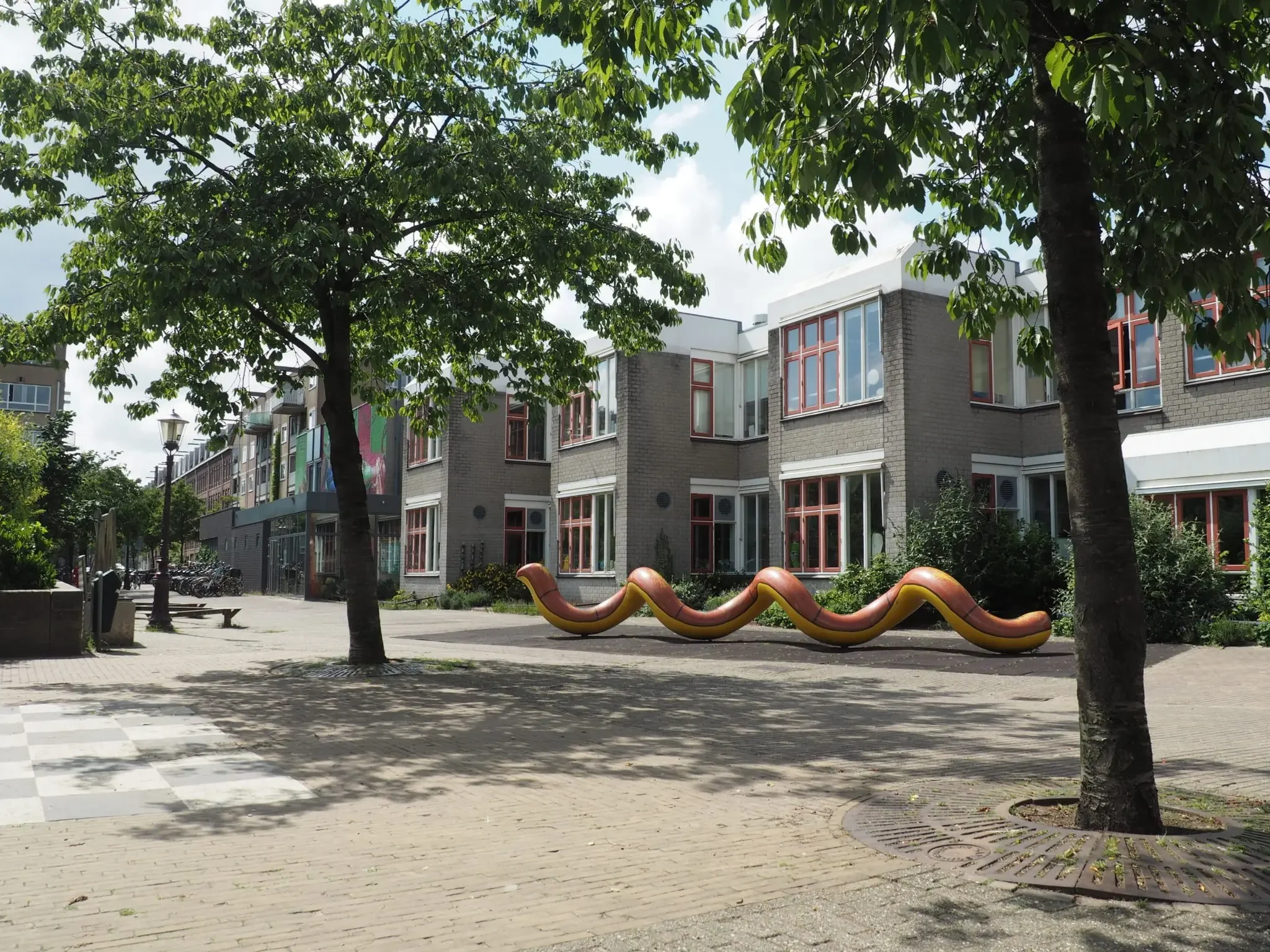 A city park with two trees, a playground slide, and a modern residential building with large windows in the background.