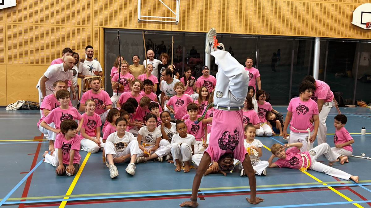 Group of children and adults in a gymnasium, some wearing pink t-shirts, with capoeira practitioners performing and posing in various positions.