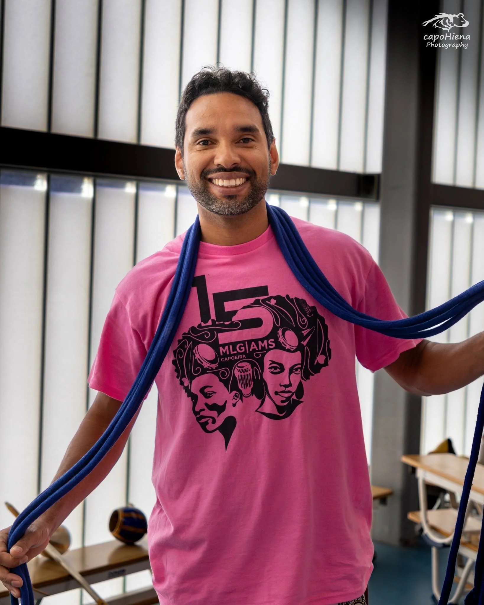 A smiling man wearing a pink T-shirt with a graphic design of three women's faces and the text 'ML GIAM'S CAPOEIRA' on it, standing indoors in front of windows with vertical blinds and holding blue ropes.
