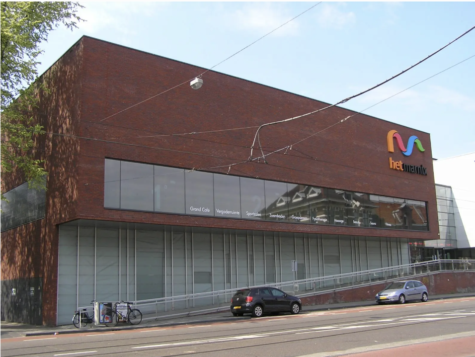 Modern brick building with large windows and colorful logo reading 'het MANIXX' on top front. Car parked in front and bicycles to the side. Overhead power lines and a tree on the left.