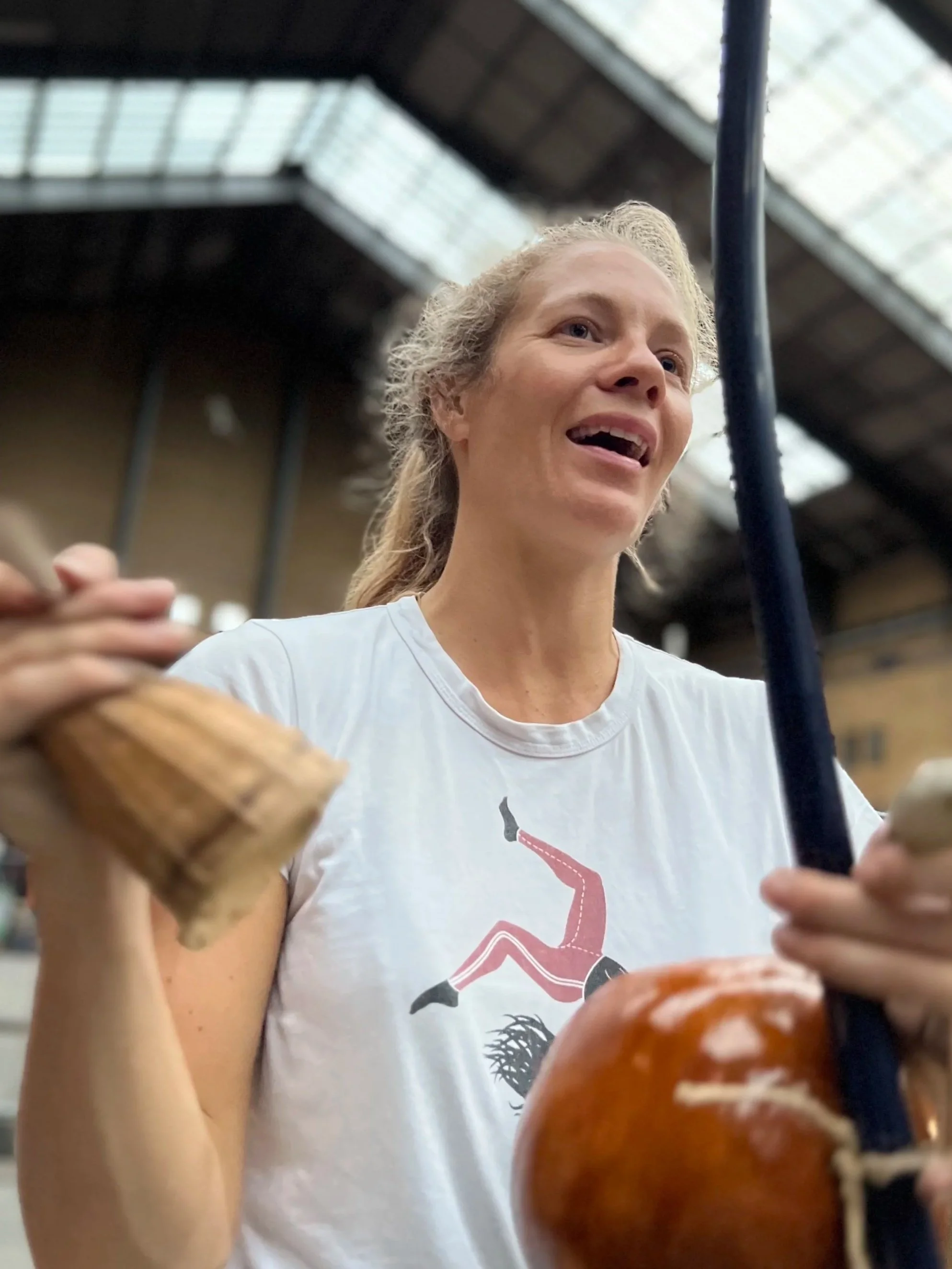 A woman with curly blonde hair smiling in a warehouse or industrial space, holding a sausage in her left hand and using her right hand to handle a cooking utensil or tool.