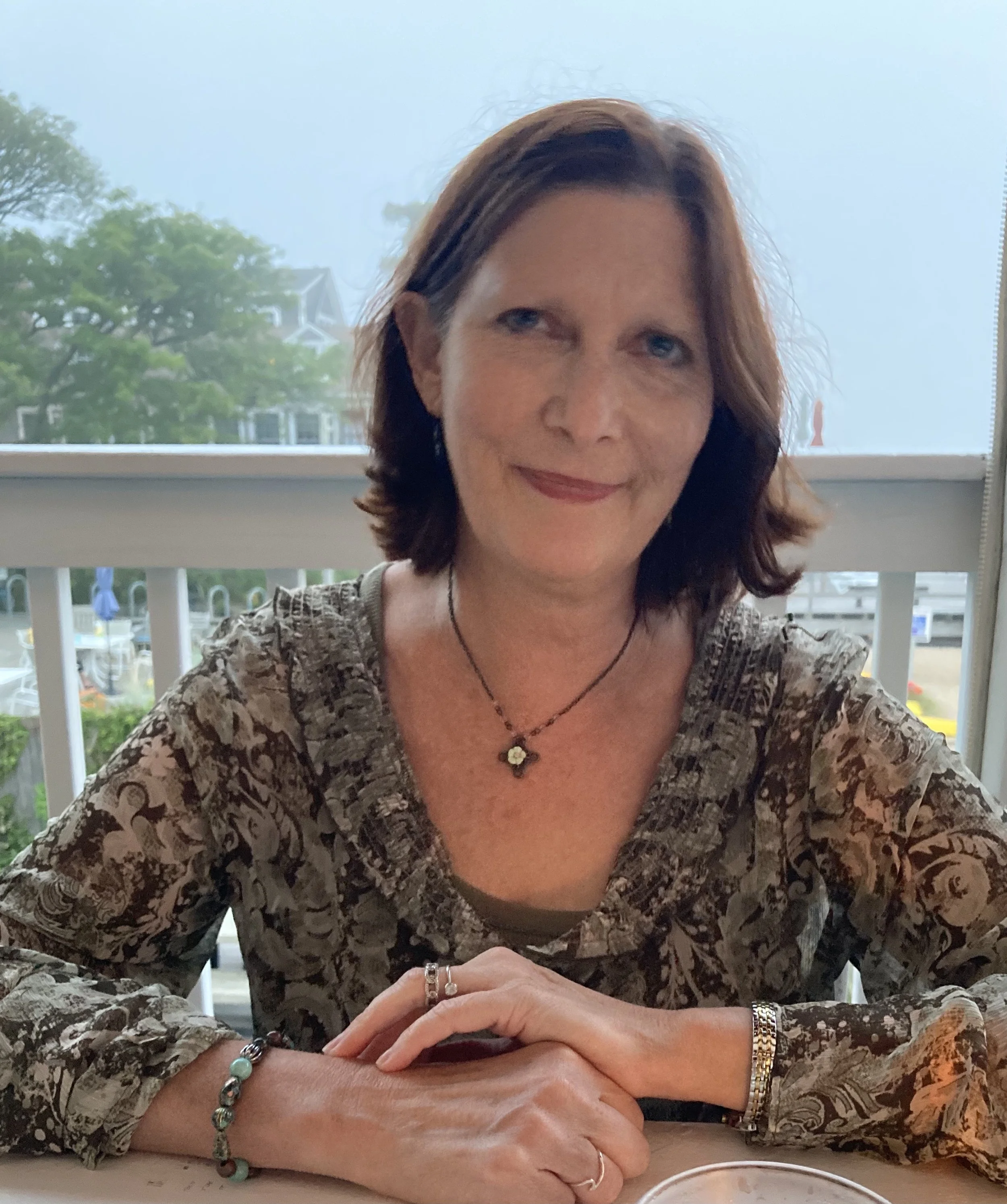 A woman with brown hair sitting at a table near a window, smiling. She is wearing a patterned top, jewelry including rings, bracelets, and a necklace. The background shows trees and houses outside the window.