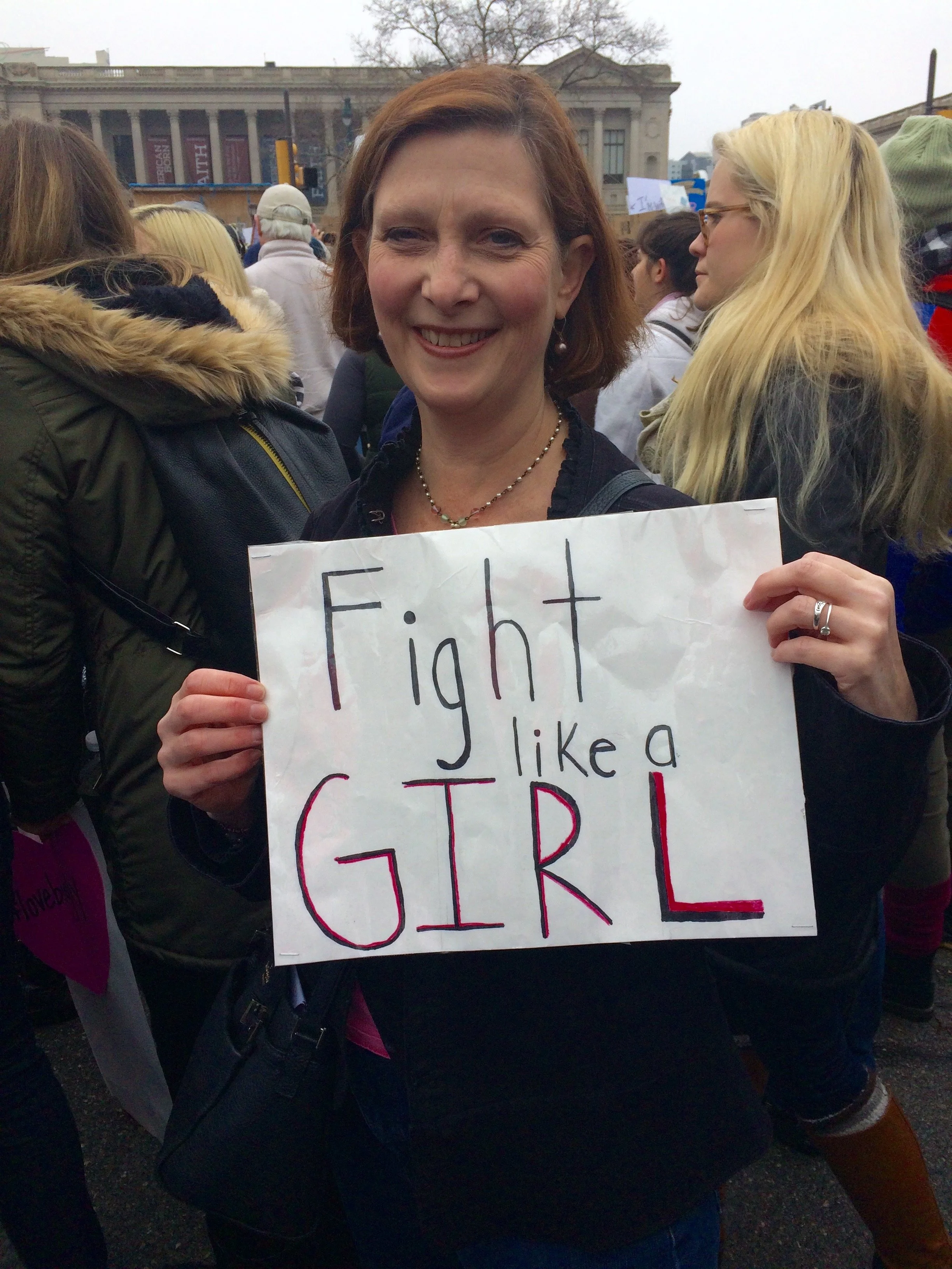 A woman at a protest holding a sign that reads 'Fight like a girl' amidst a crowd of protesters.