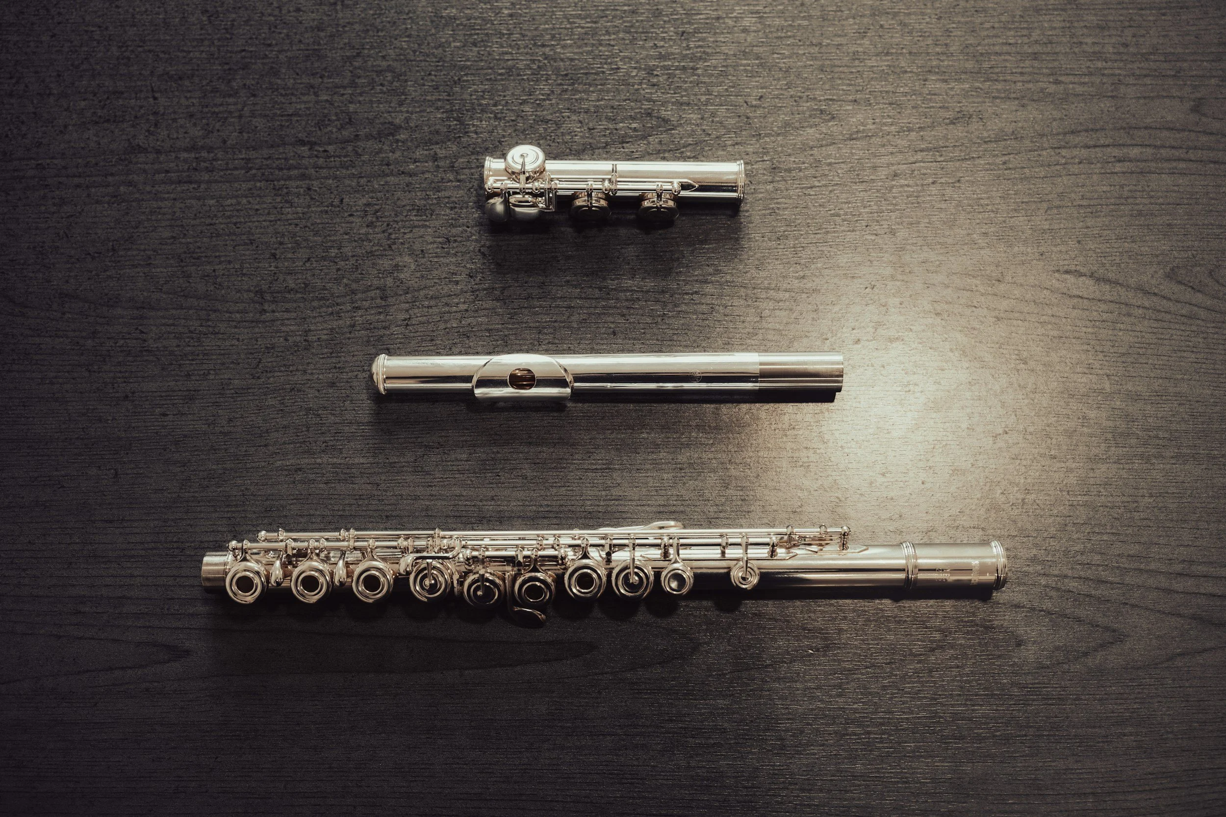 Four silver musical instruments arranged horizontally on a dark wooden surface: a piccolo, a flute, an oboe, and a clarinet, from top to bottom.