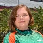 A person with shoulder-length brown hair and glasses standing outdoors in front of green plants, wearing a teal and orange sports jersey.
