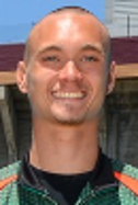 A smiling man with a shaved head, wearing a black shirt and turquoise necklace, outdoors with a building in the background.